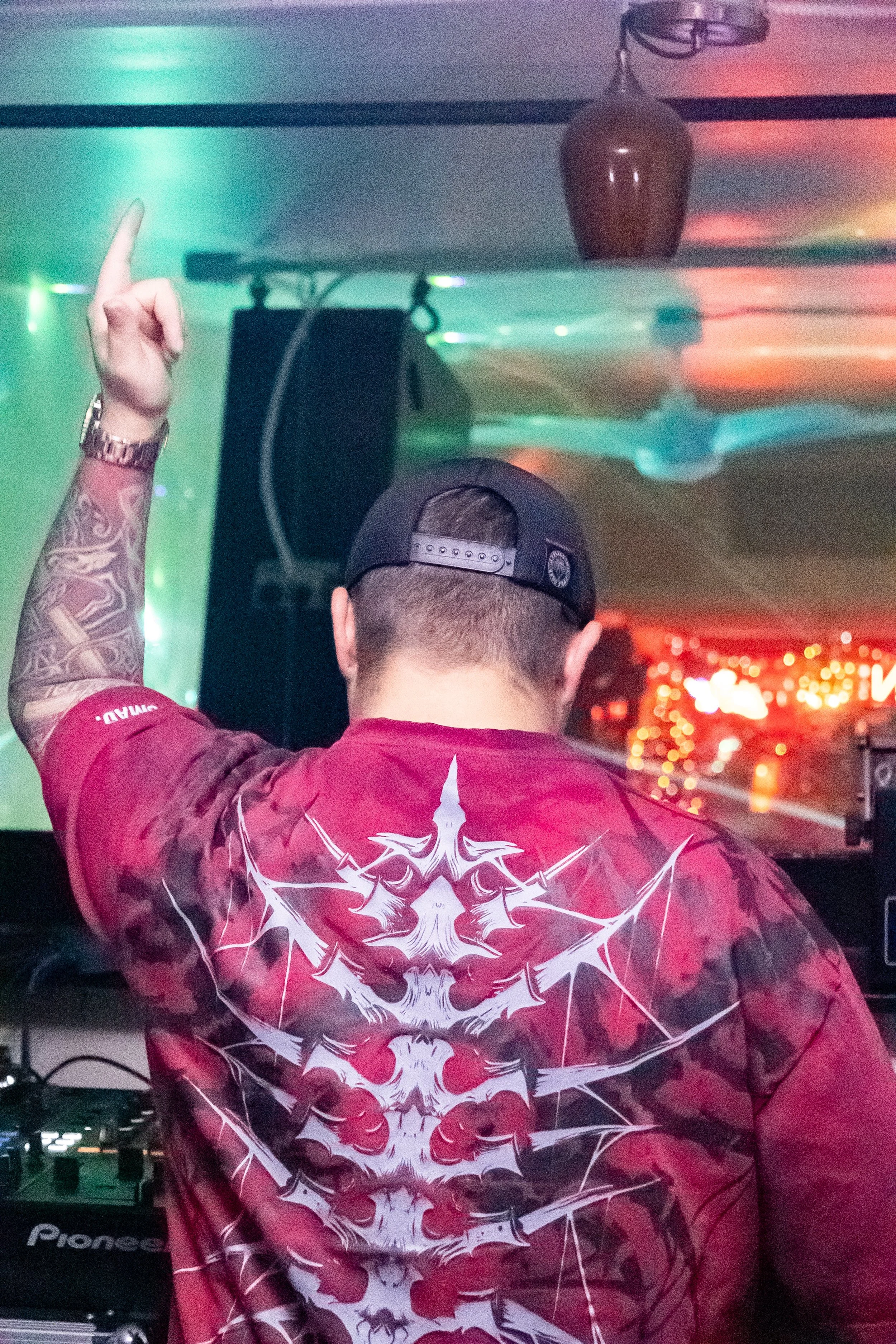 A DJ wearing a black baseball cap and a red graphic T-shirt with white designs, standing behind DJ equipment, with his arm raised in a club with colorful lighting and a ceiling fan.
