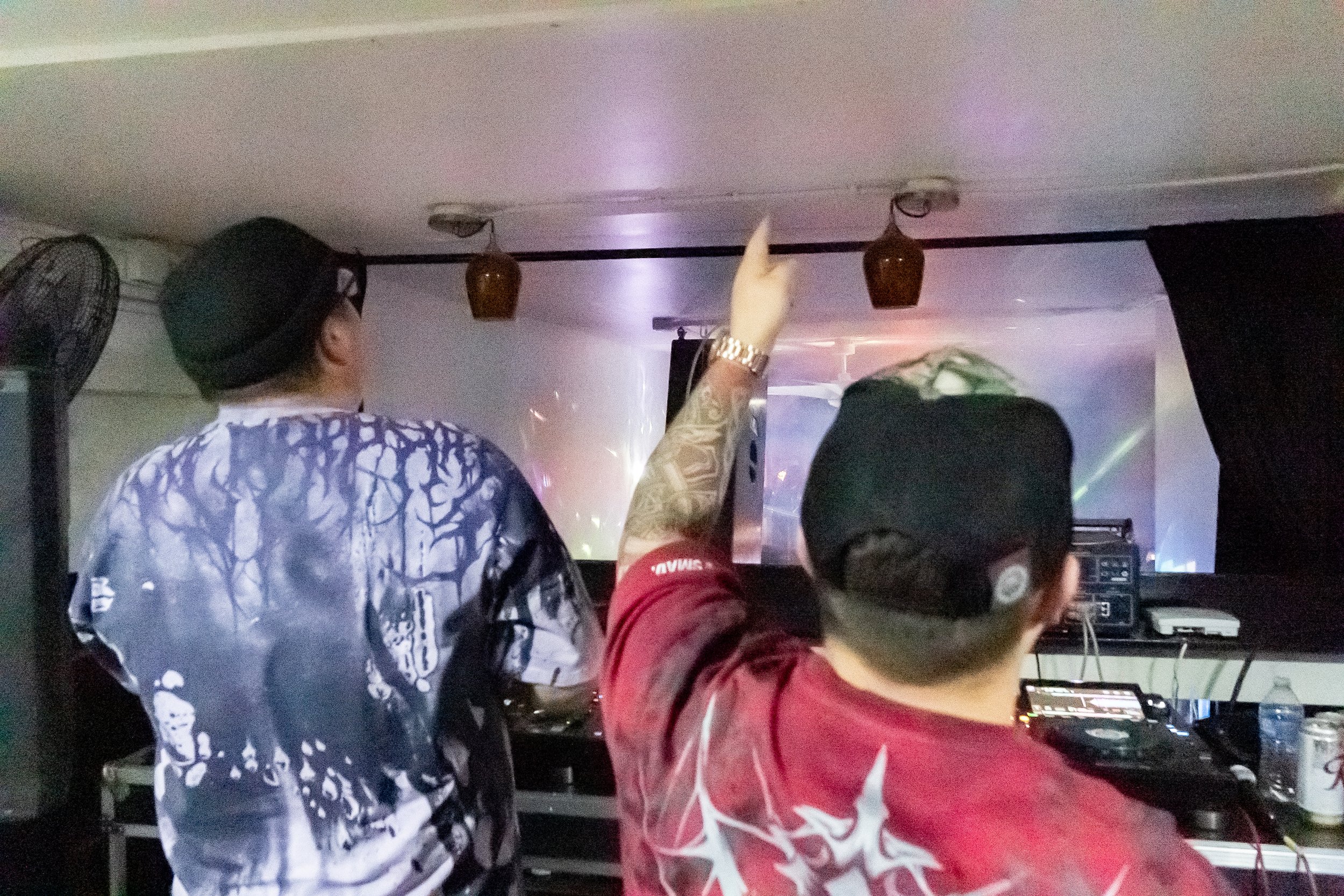 Two men are in a dark room with electronic equipment, one is facing away, wearing a black cap and a red shirt with white graphic, the other is facing away, wearing a black hat and a shirt with a black and white pattern, pointing towards the ceiling w