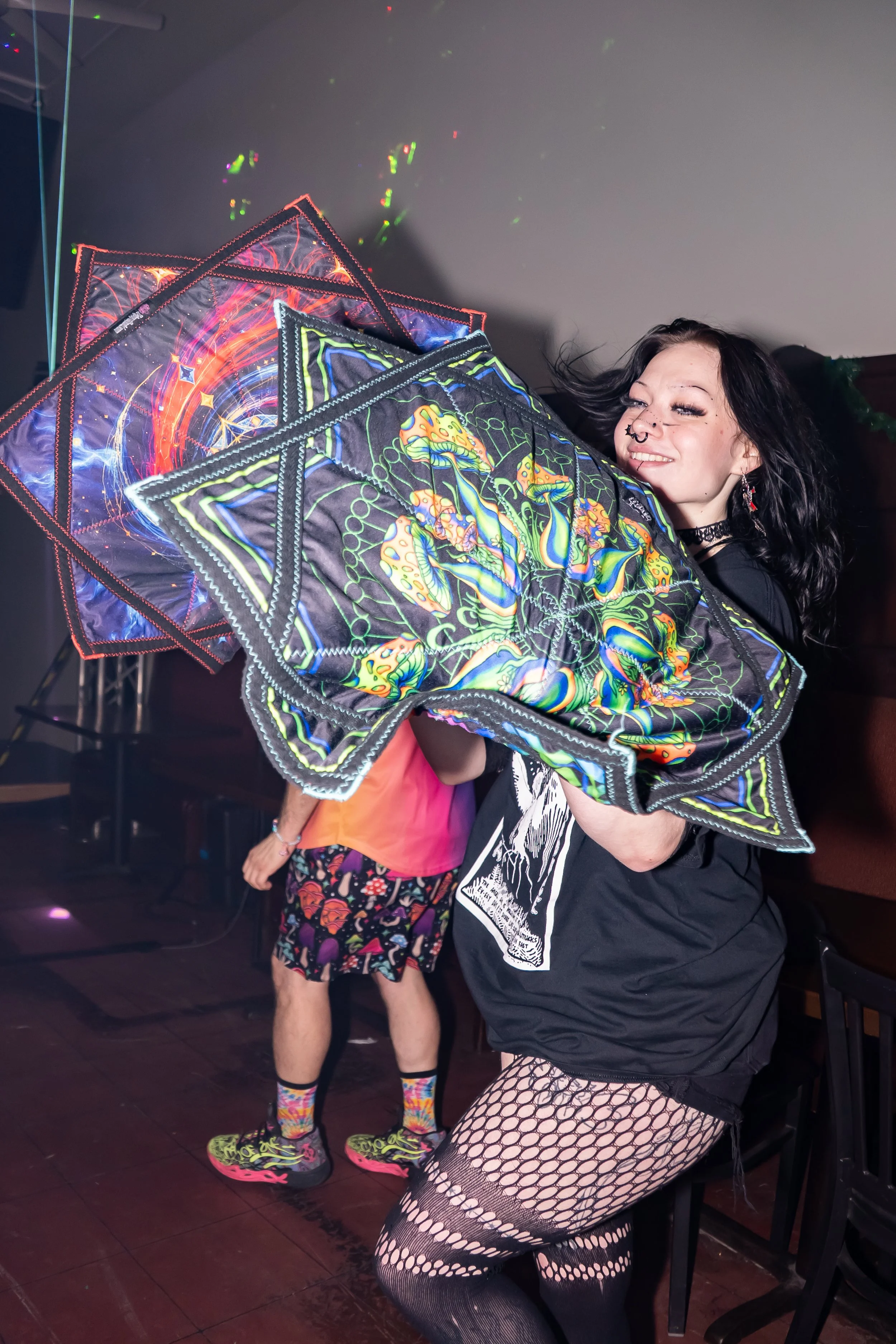 A person with black hair, black fishnet stockings, and a black outfit holding LED-lit, colorful kites at a party or event.