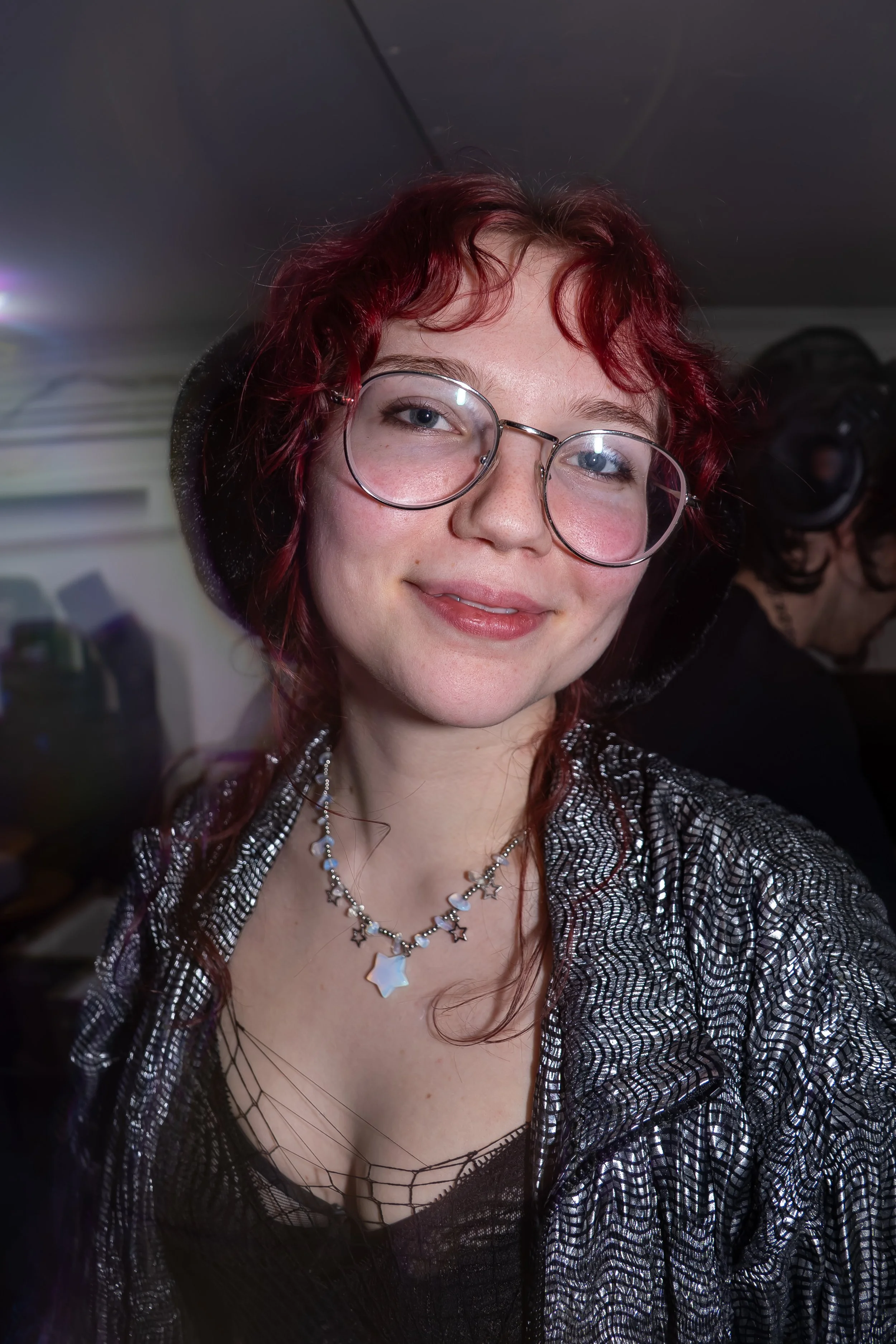 A young woman with red hair, glasses, and a silver star necklace taking a selfie at a party or event.