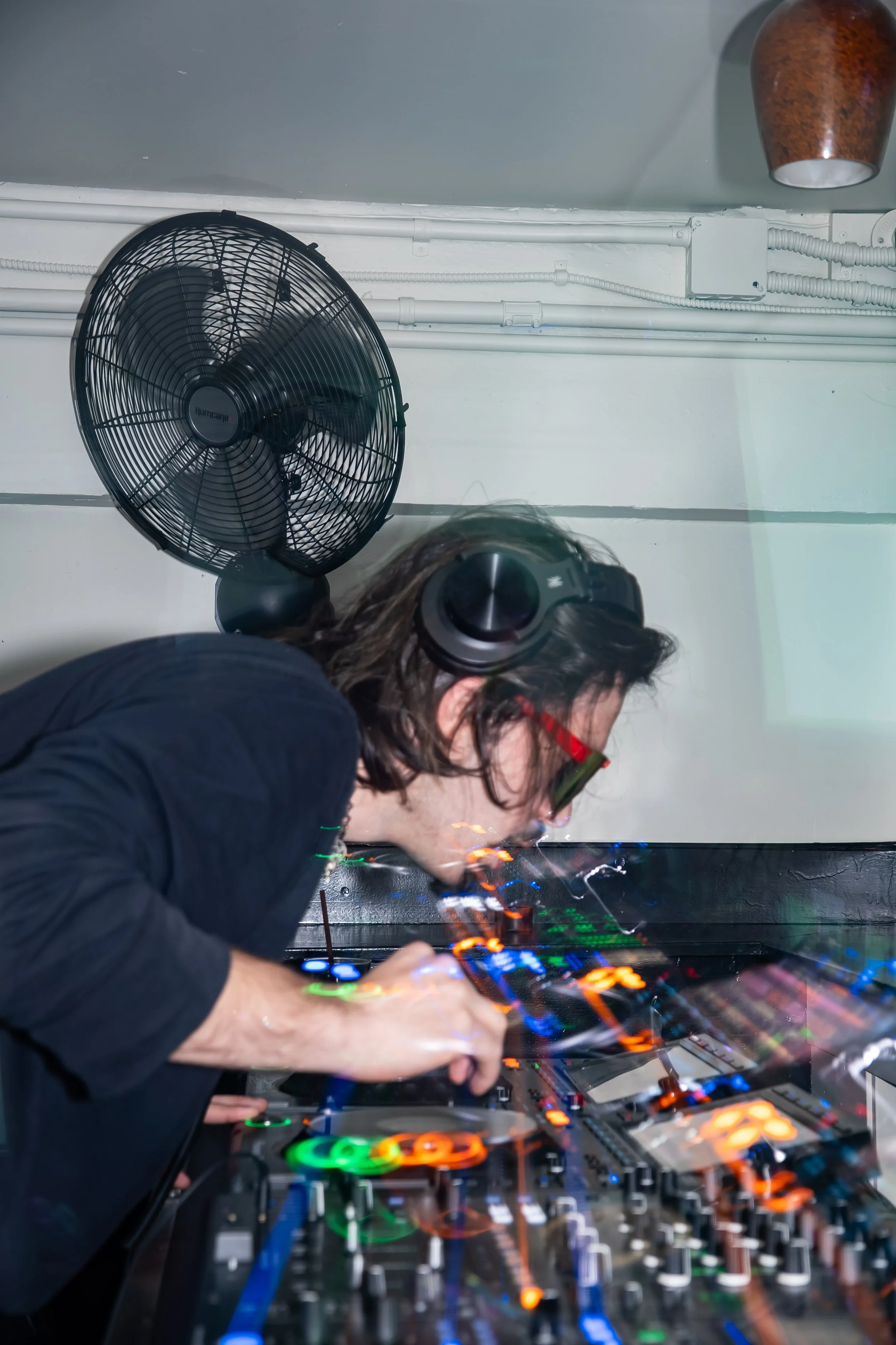 A DJ with long hair, red glasses, wearing large black headphones, is playing music on a DJ mixer with colorful LED lights. There is a large black fan mounted on the wall in the background.