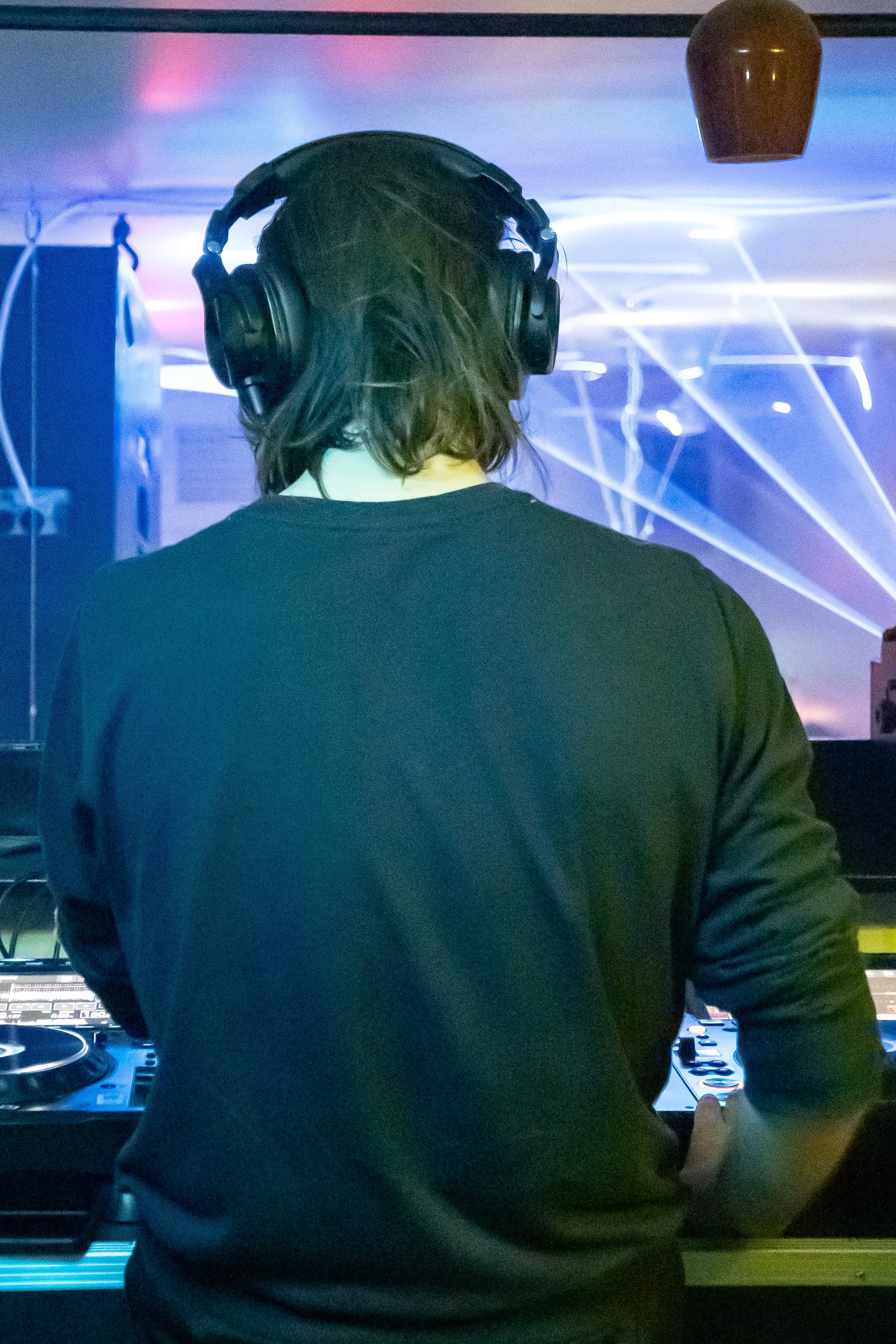 DJ with long hair wearing headphones in front of a DJ console with colorful lighting effects in the background.