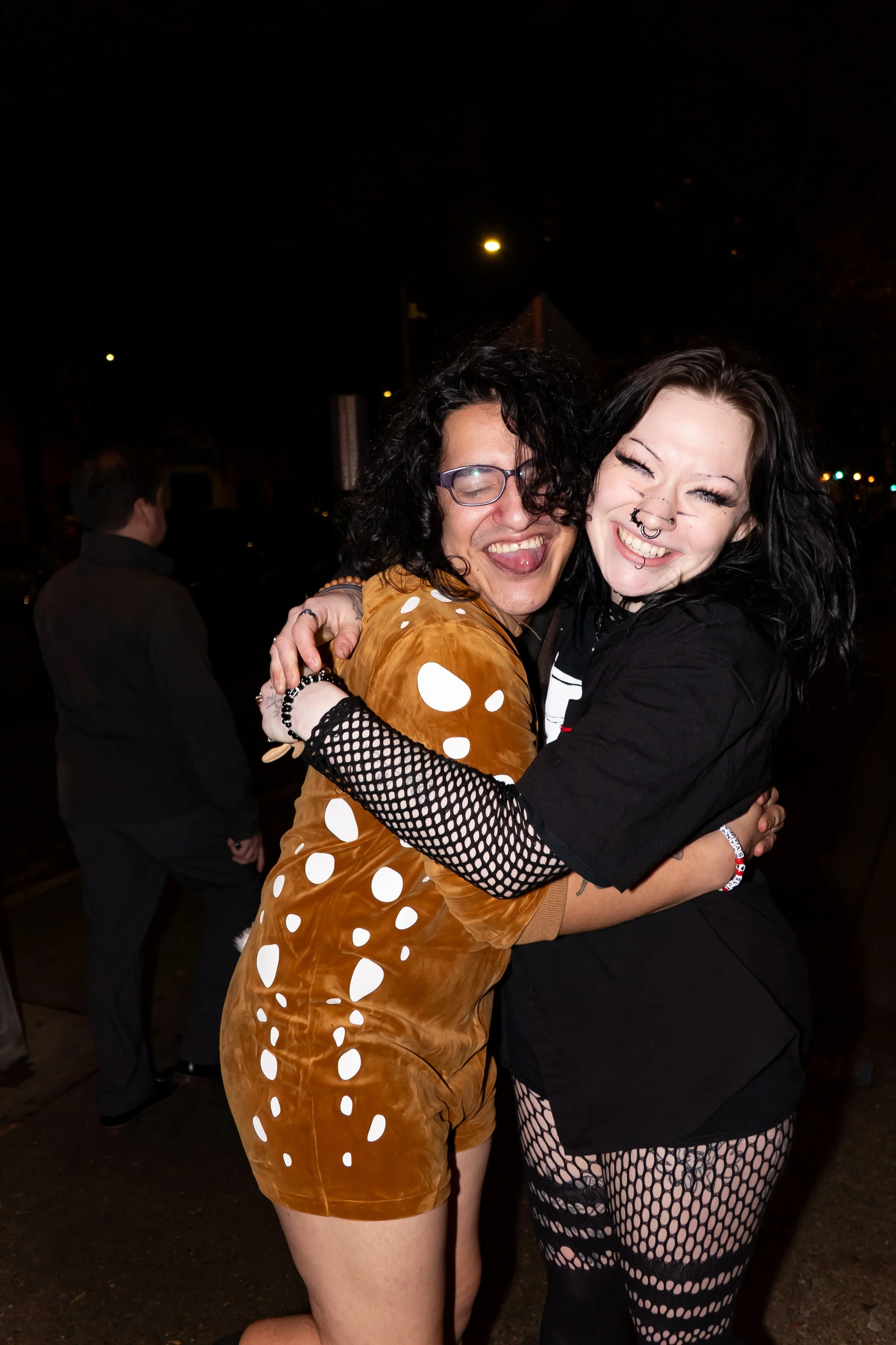Two people hugging and smiling at night, dressed in costumes; one wearing a brown and white polka dot outfit, the other in black fishnet clothing.