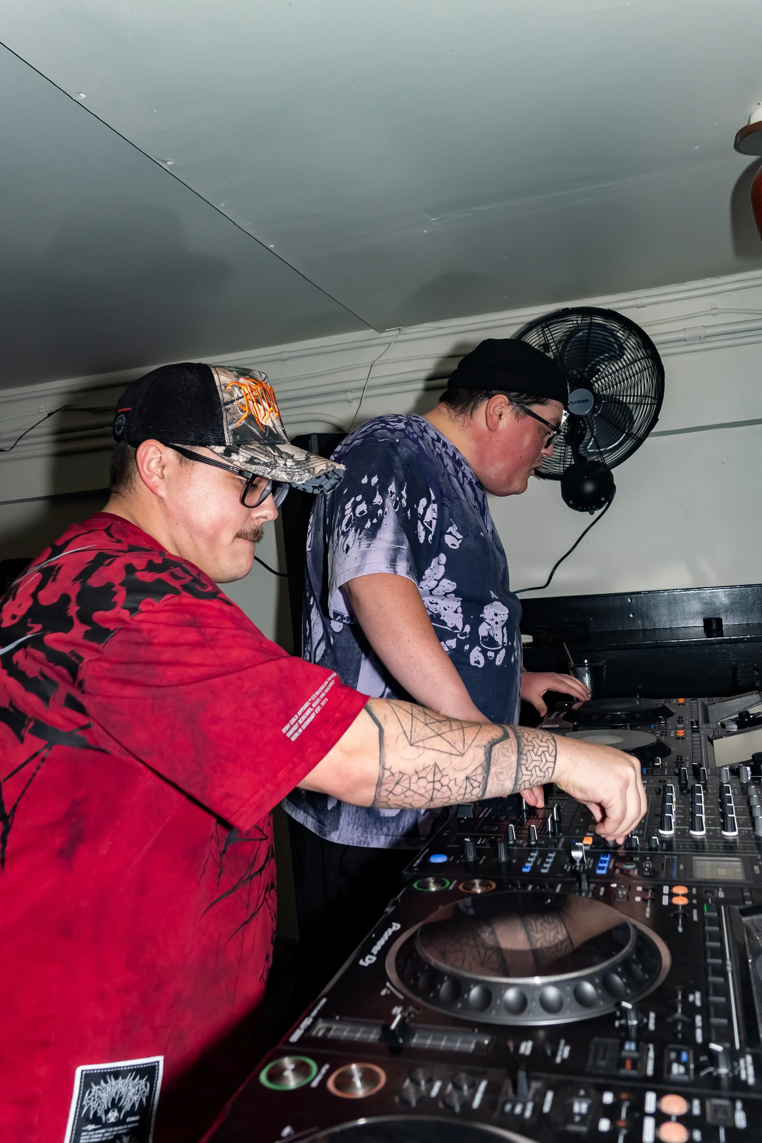 Two men DJing with equipment in a room, one wearing a red tie-dye shirt and a black baseball cap, the other in a black beanie and dark tie-dye shirt, with a wall fan in the background.