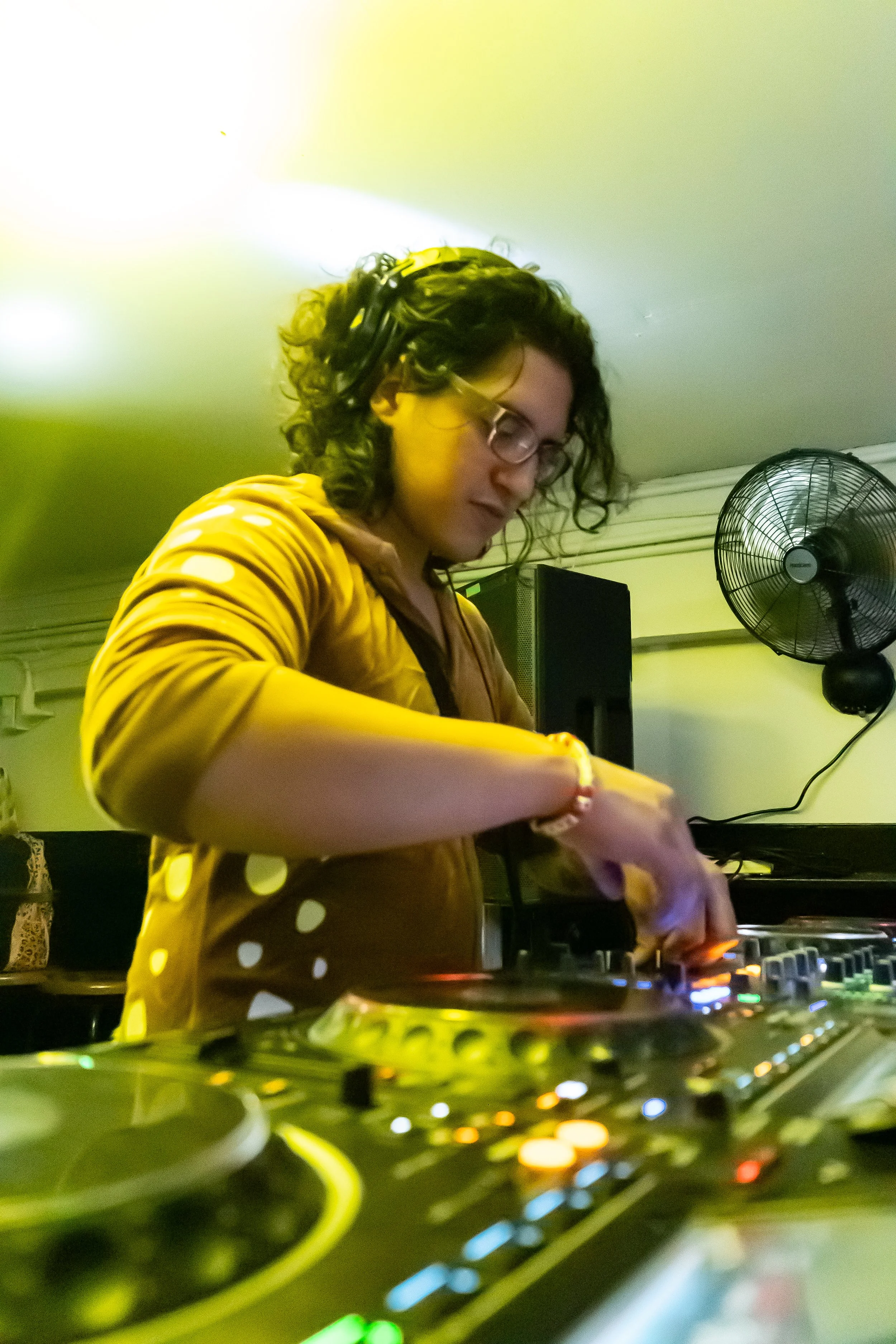 A female DJ with curly hair and glasses, wearing a yellow polka dot jacket, playing music on a DJ mixer in a softly lit room.