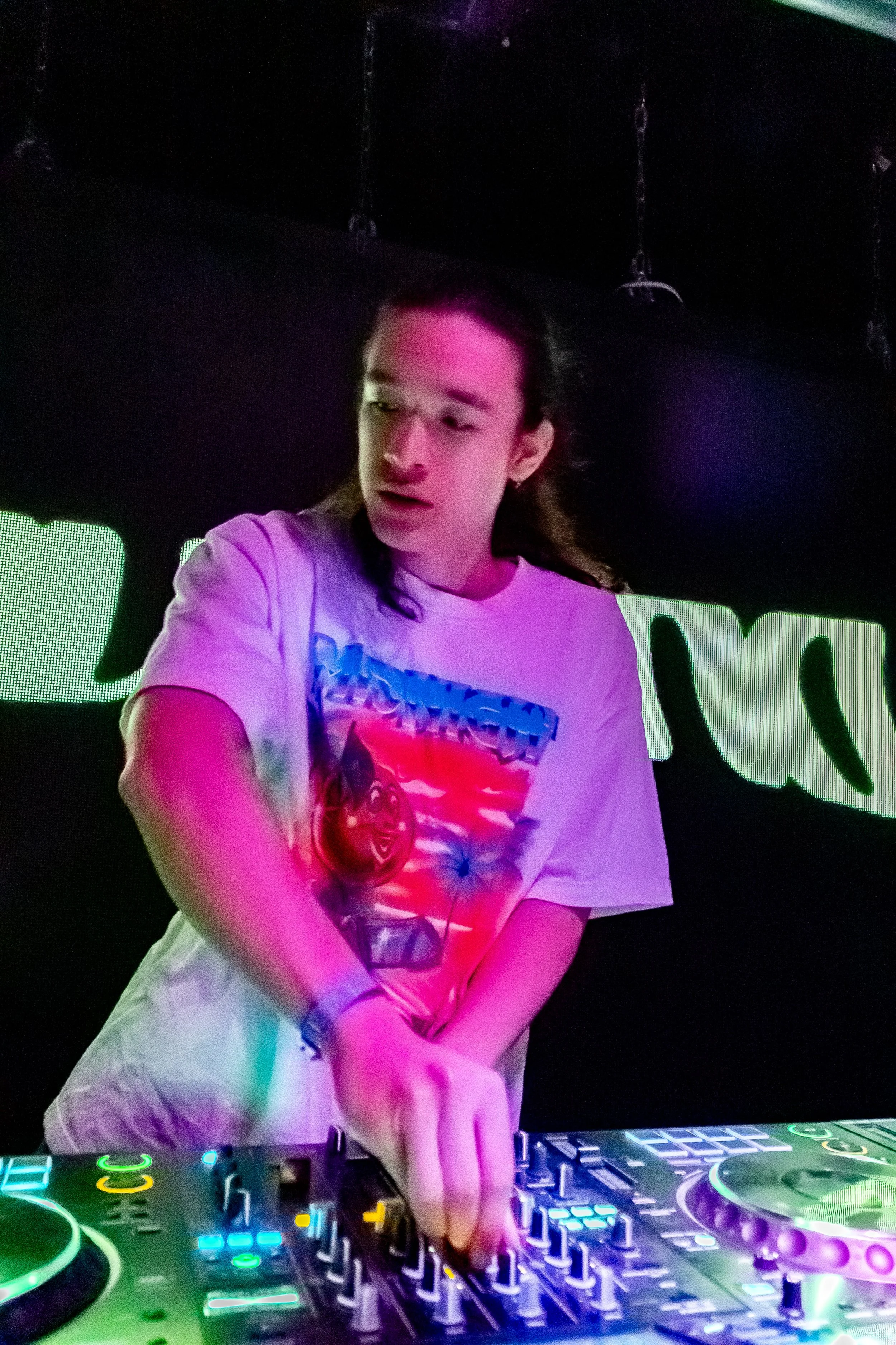 Person DJing at a club with illuminated equipment and a digital screen in the background.