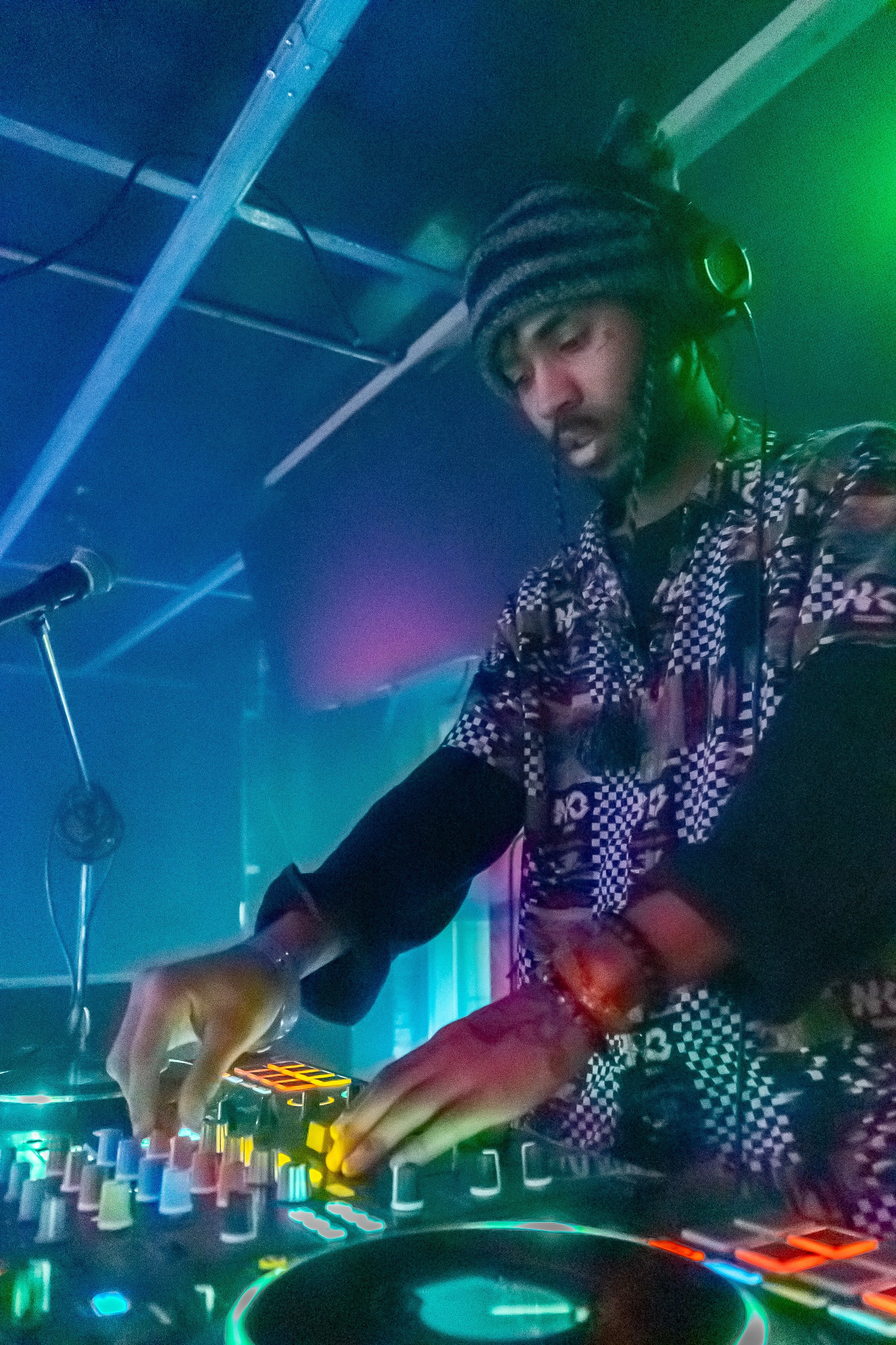 A DJ wearing a checkered shirt and headphones, using a DJ mixer in a club with colorful lighting.