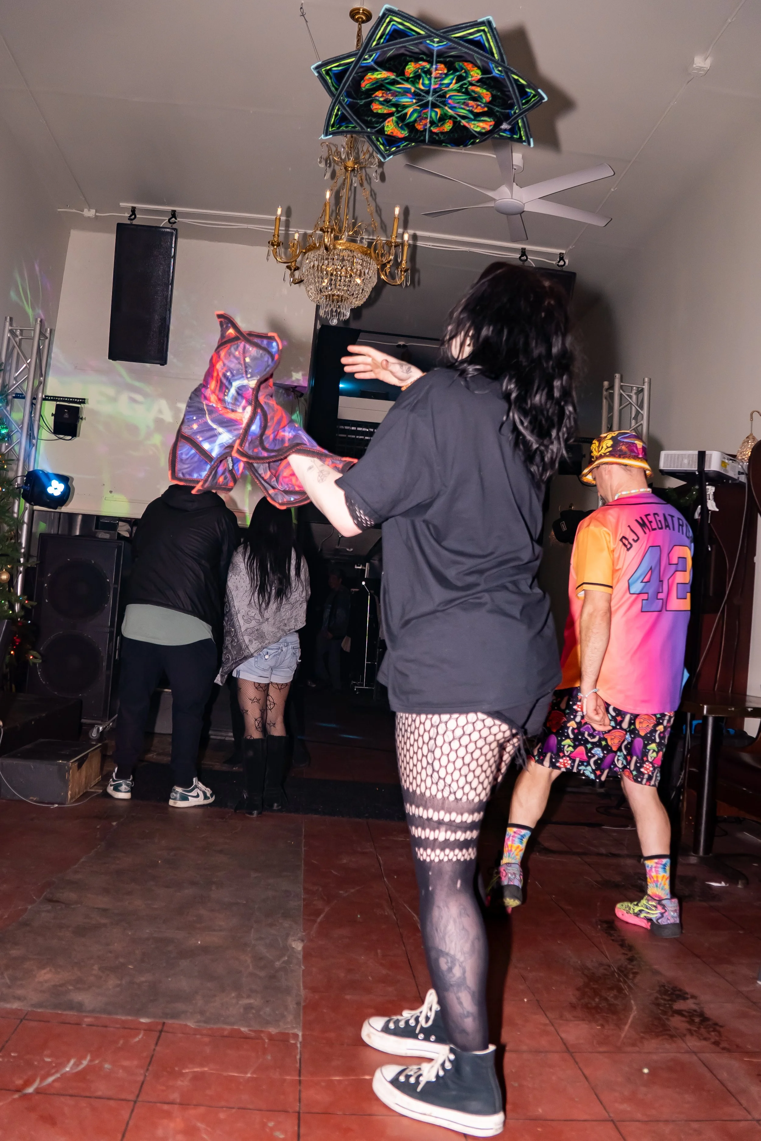 People dancing at a party or club with colorful costumes and lighting, a chandelier, and sound equipment.