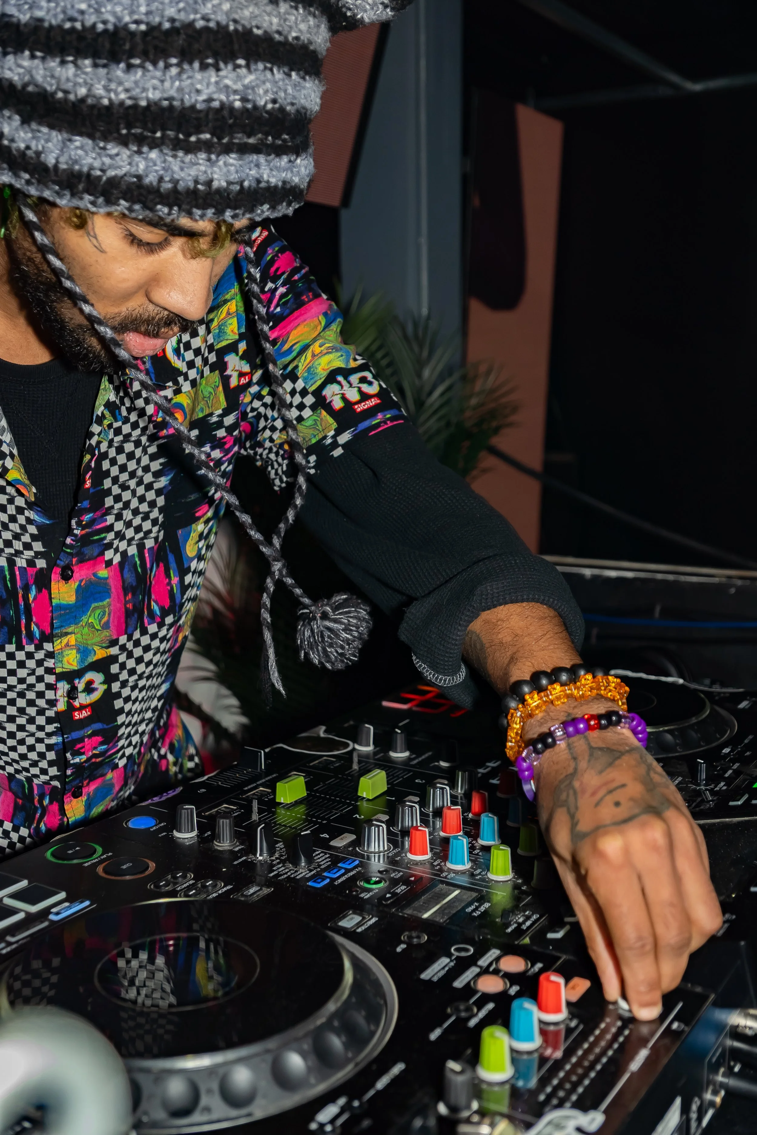 A DJ wearing a colorful patterned shirt and a knit hat, adjusting controls on a DJ mixer with various colorful knobs and sliders.