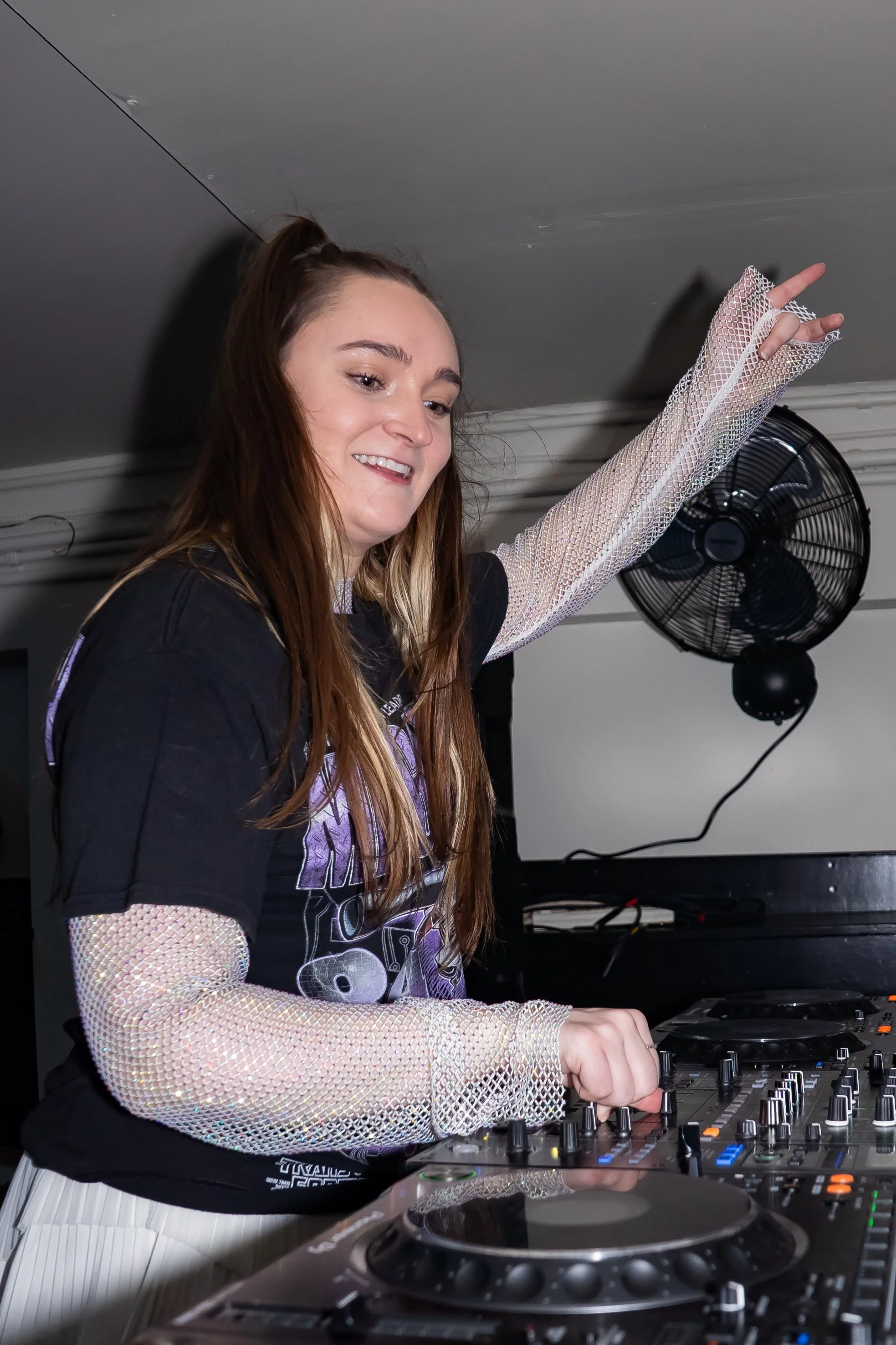 A young woman DJing at a party, wearing a black t-shirt with purple graphics, mesh long sleeve shirt underneath, and smiling while operating DJ equipment.