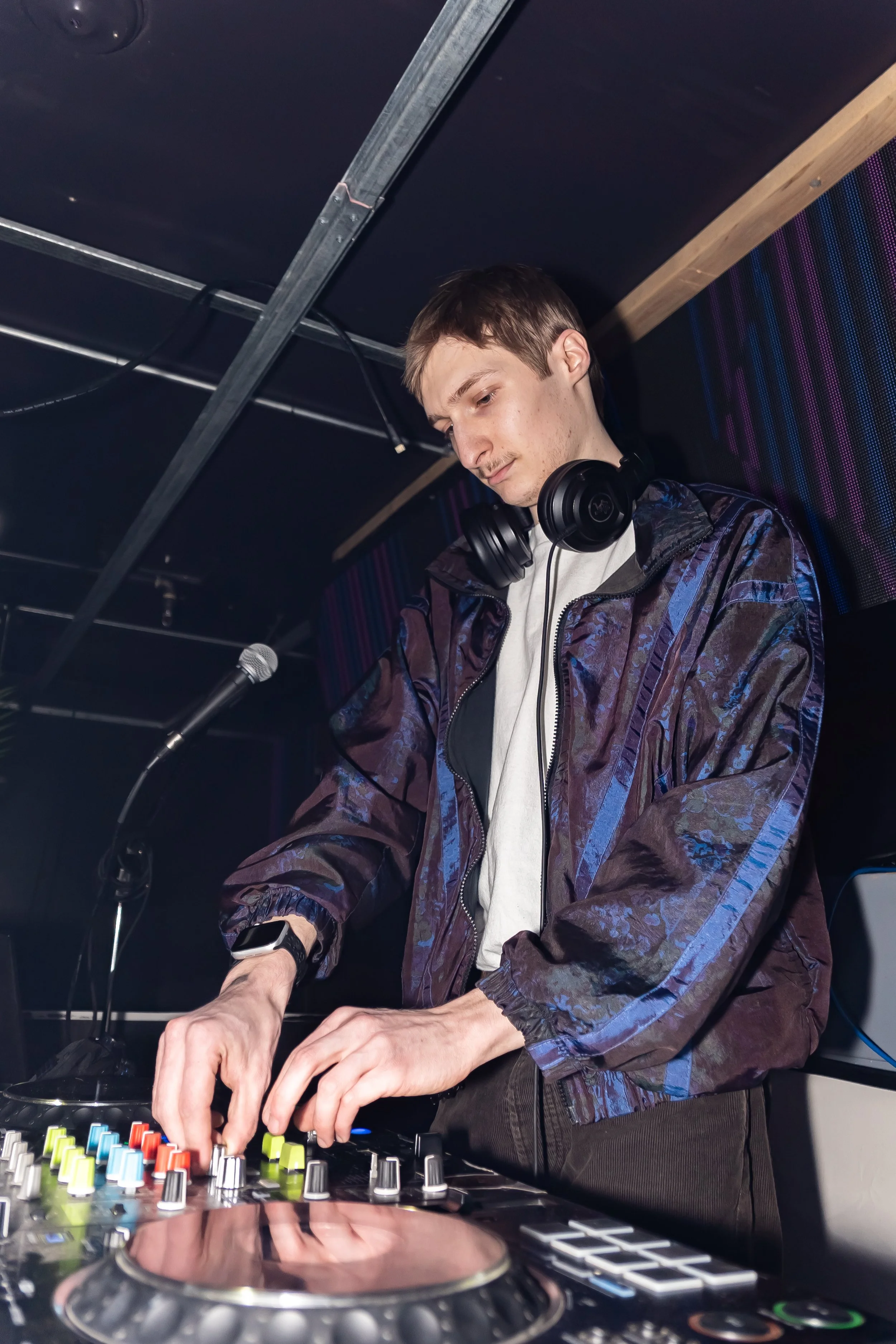 A young male DJ wearing a headset, operating a DJ mixer with colorful knobs and buttons in a dimly lit nightclub or music venue.