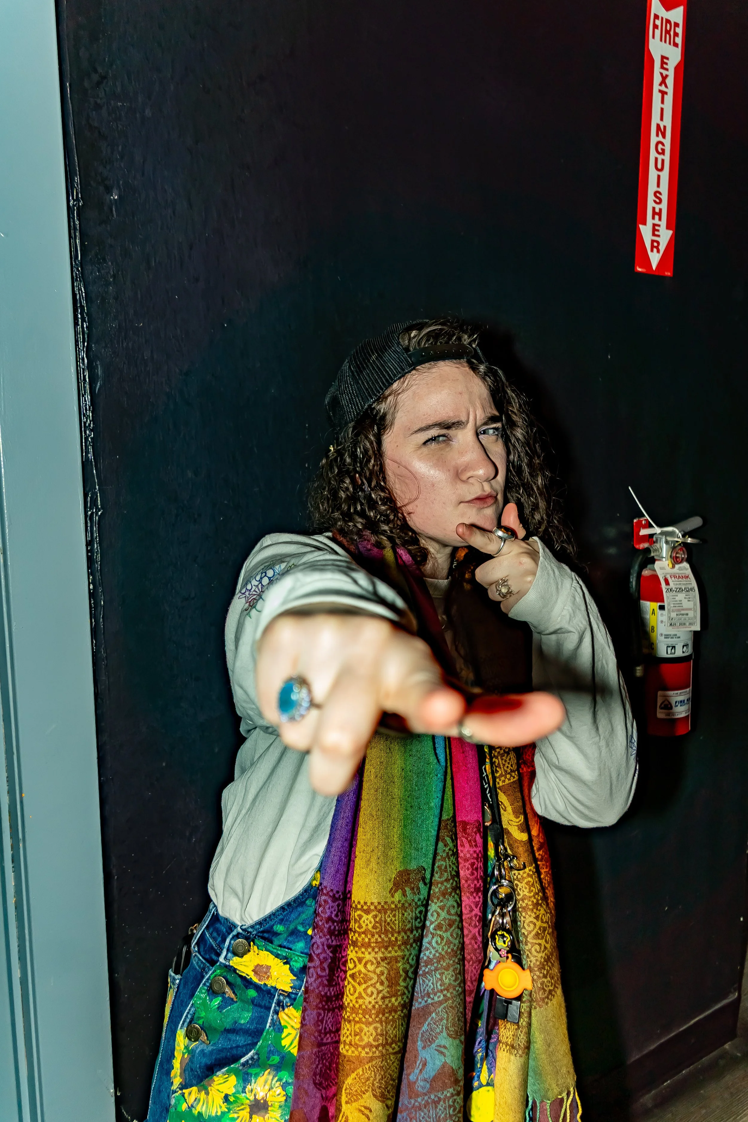 Young woman pointing at the camera with a serious expression, wearing a colorful scarf, a white jacket, and a backward baseball cap, standing near a fire extinguisher on a dark wall.