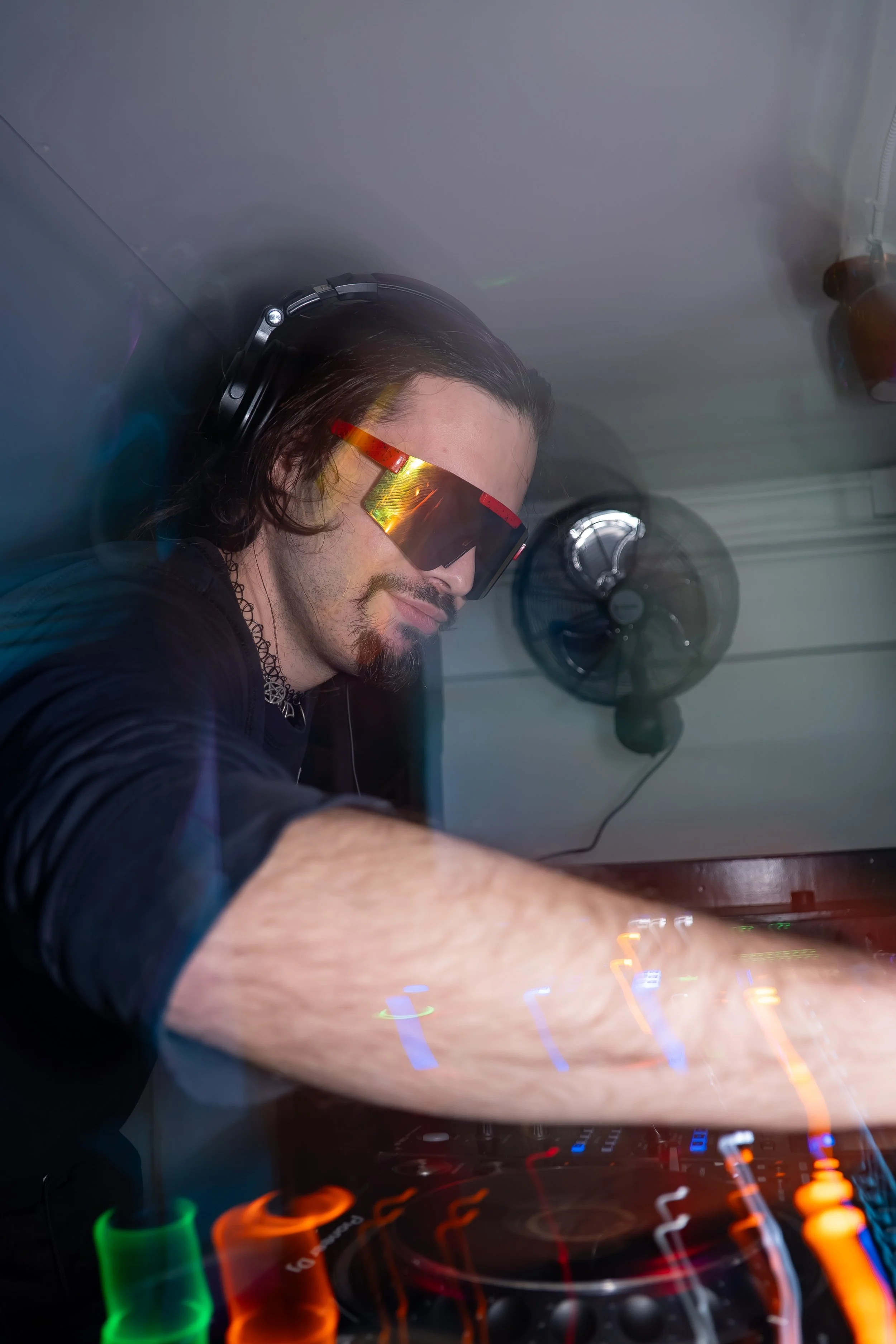 A male DJ wearing reflective sunglasses, headphones, and a black shirt is mixing music at a club or party, with colorful light trails and a fan in the background.