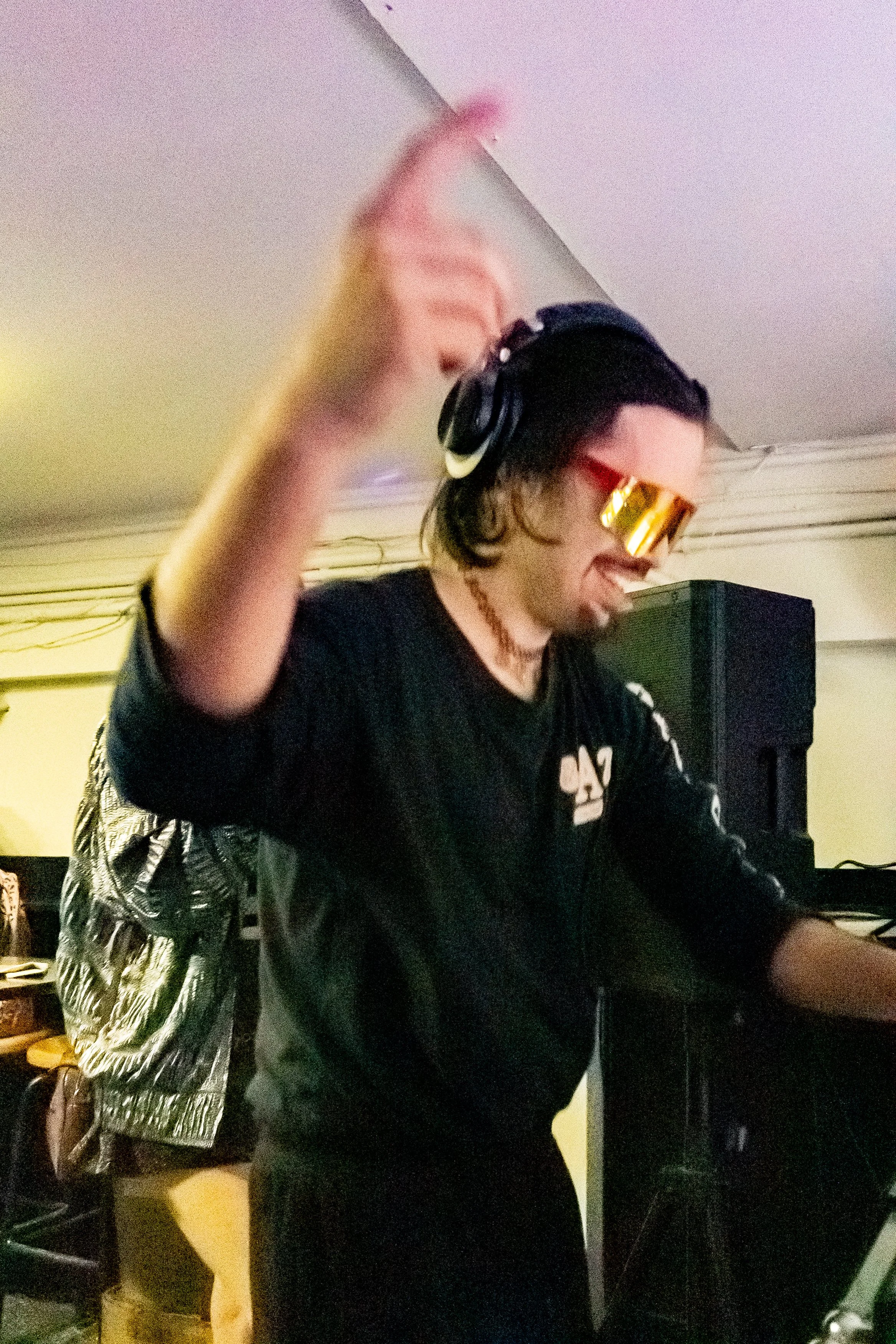 Person with dark hair wearing gold sunglasses and headphones, DJing at a music event.