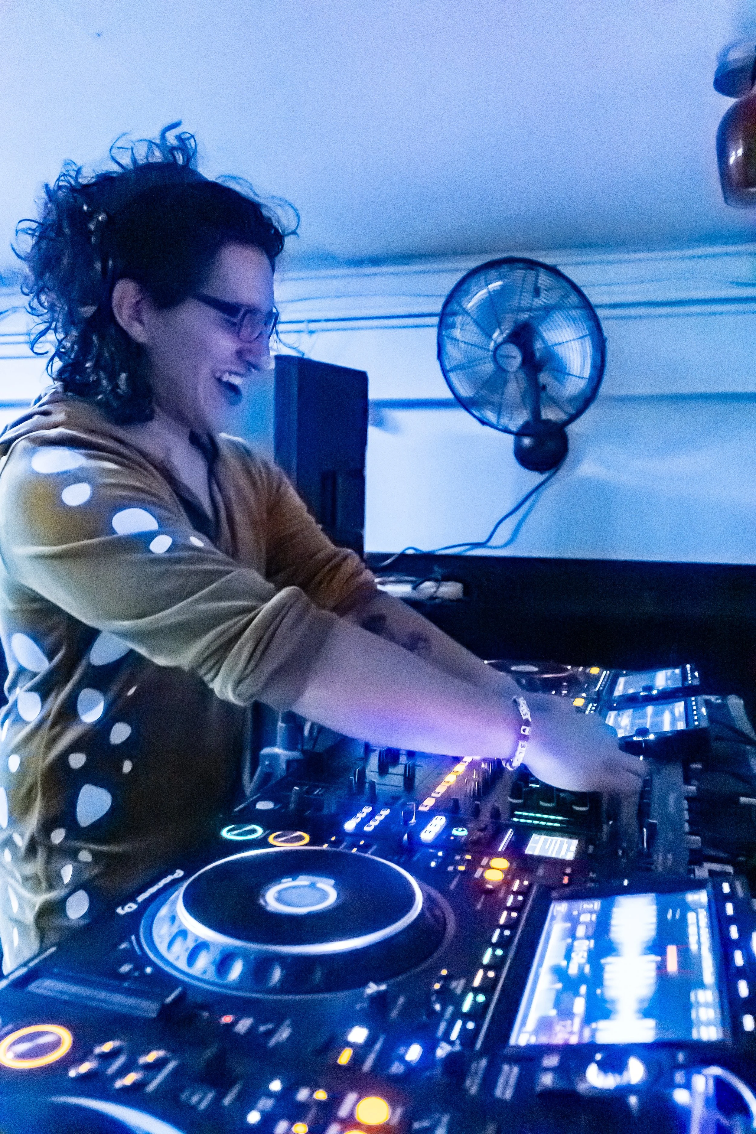 A man with curly hair and glasses DJing with a smile in a blue-lit room, wearing a polka dot top, behind a professional DJ controller.