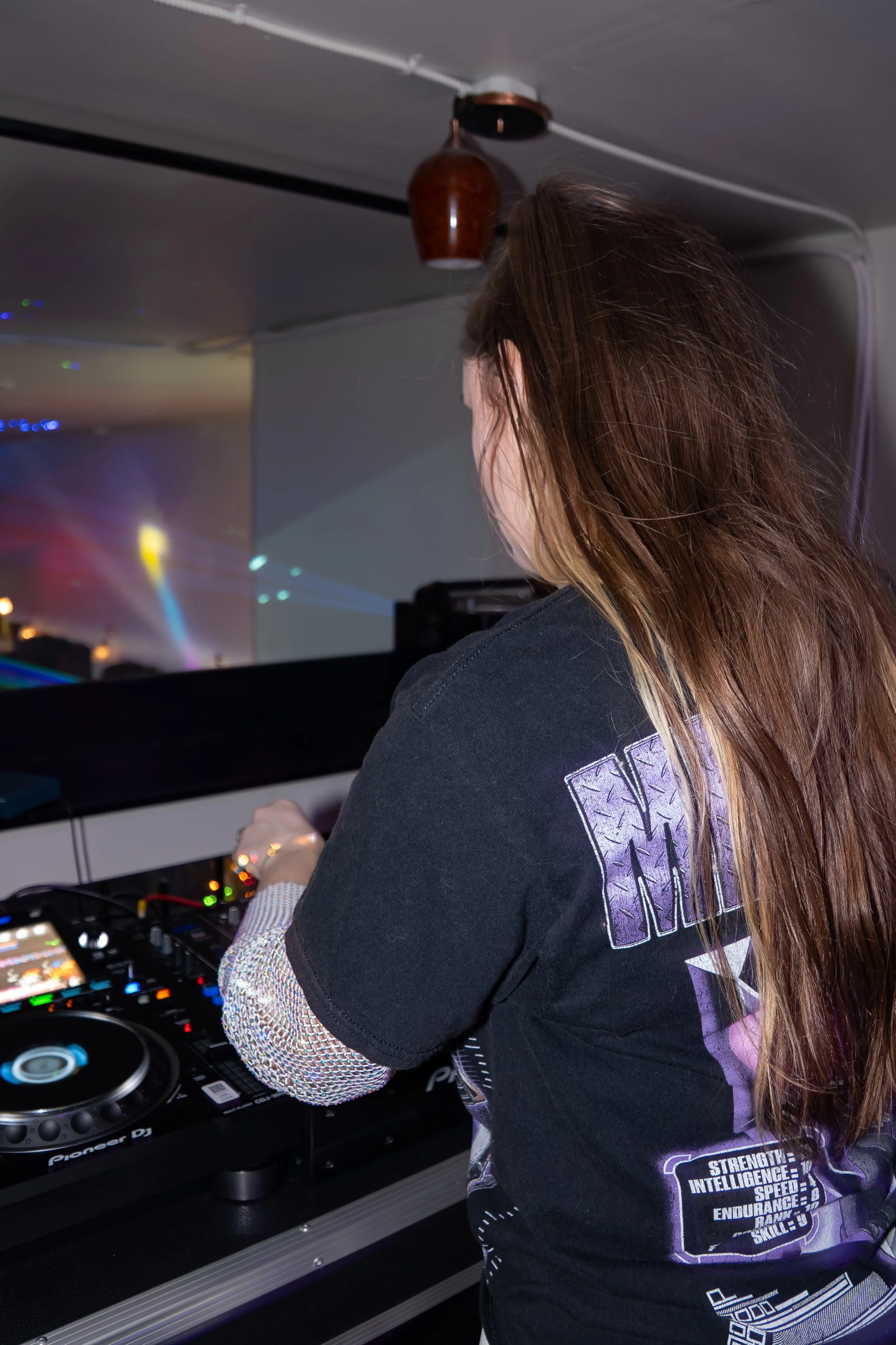 A woman with long brown hair DJing at a club or party, wearing a black t-shirt with purple and white designs, and mixing music on DJ equipment.