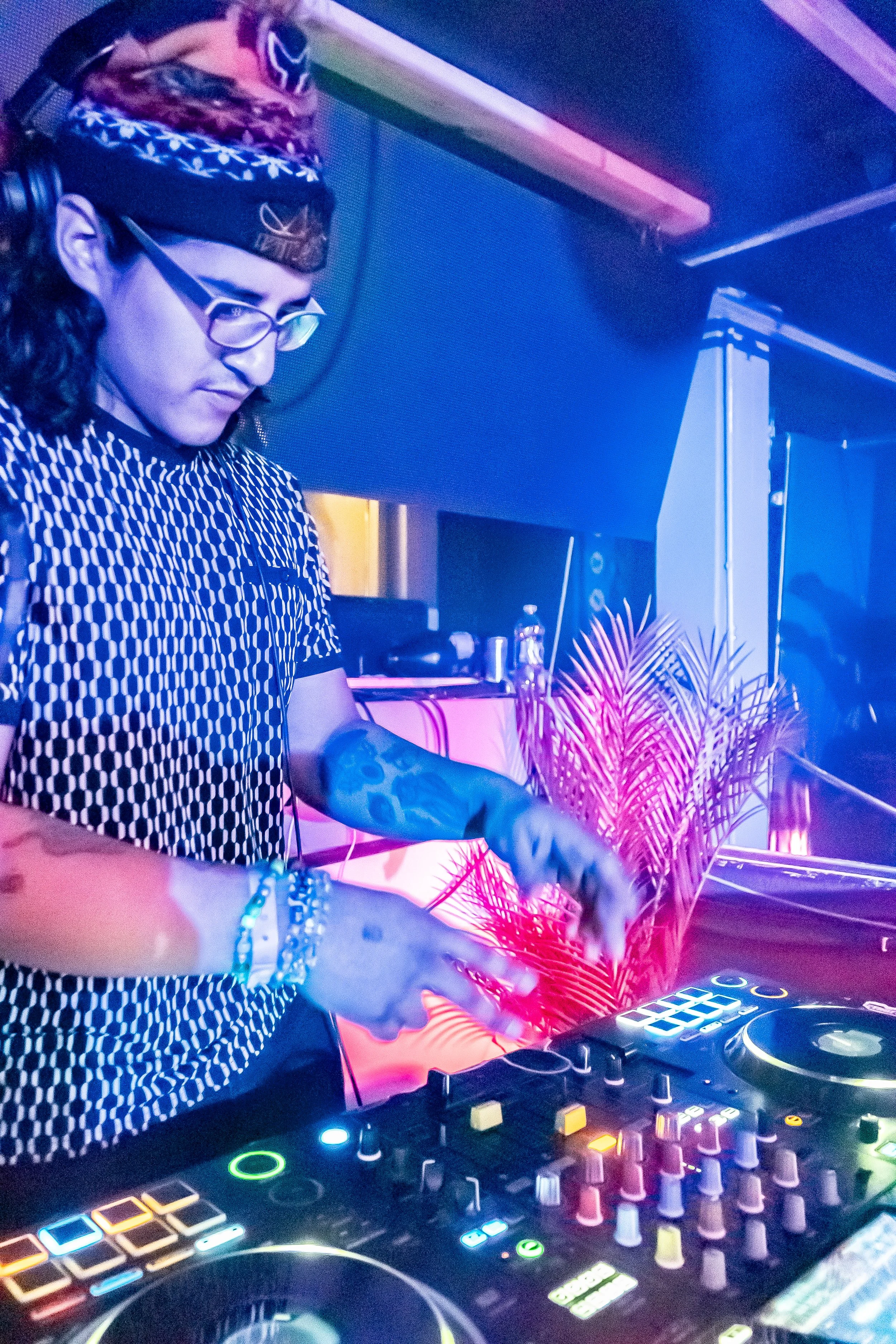 A DJ wearing glasses, a patterned shirt, and a bandana, operating DJ equipment with colorful lights and a pink plant in the background.