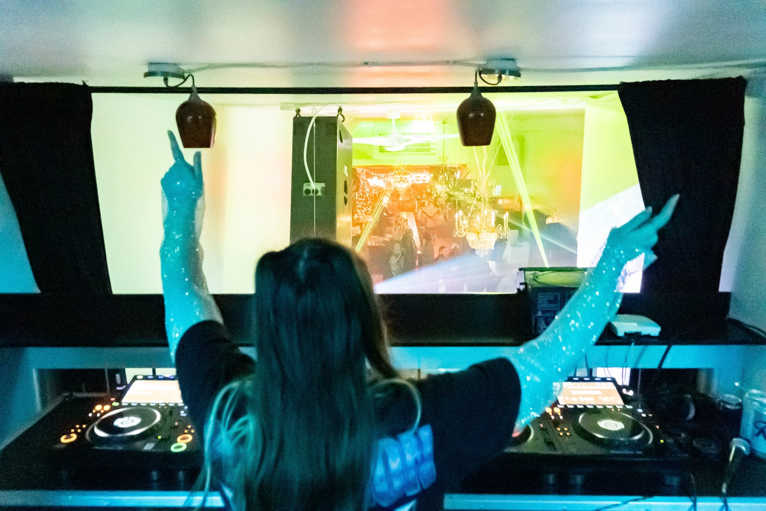 A DJ woman with glittery blue arm sleeves performing at a club with colorful lights and a large screen in front of her, pointing towards the crowd.