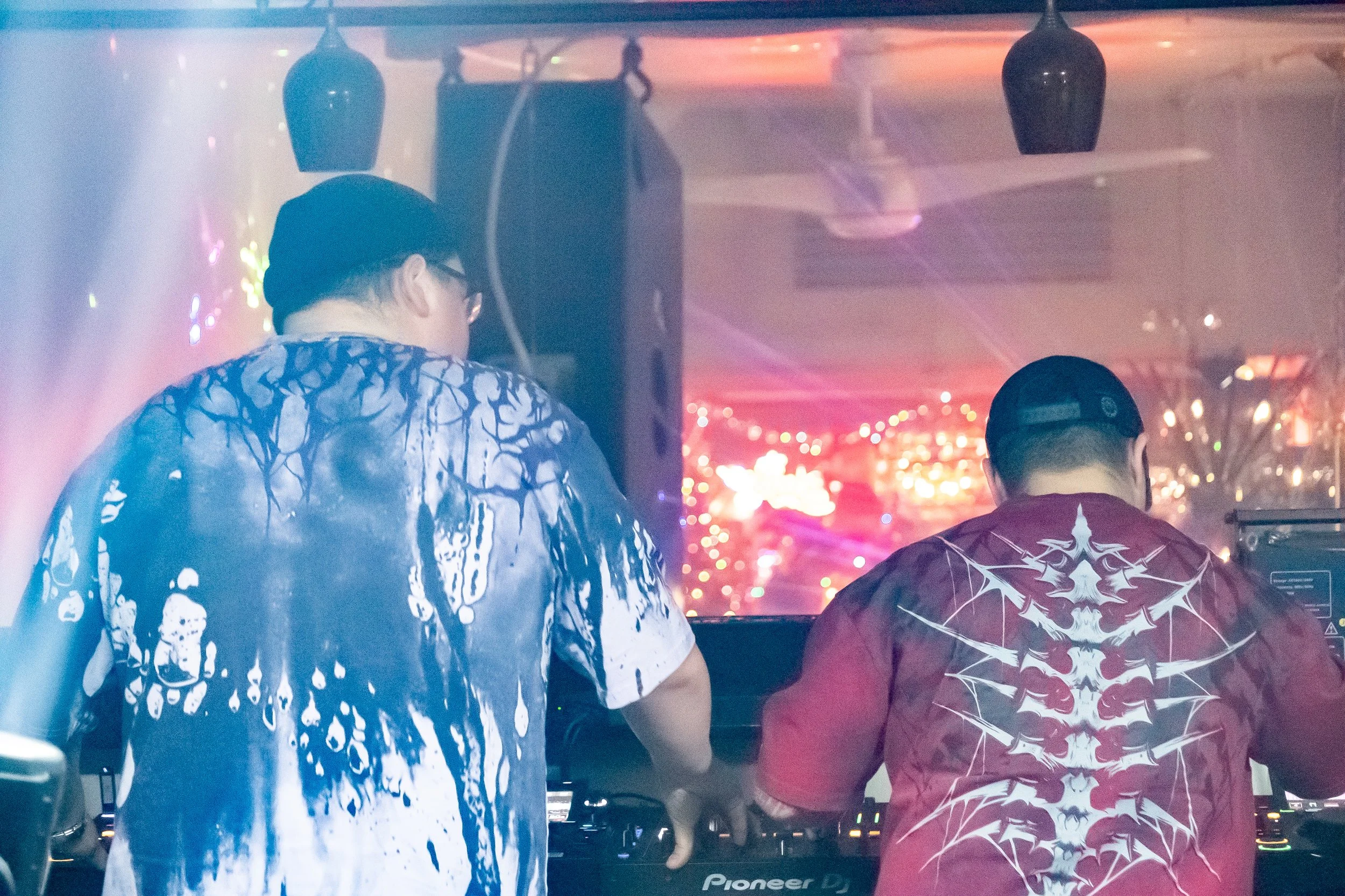 Two DJs wearing graphic T-shirts and caps are performing at a nightclub with colorful lights and decorations in the background.