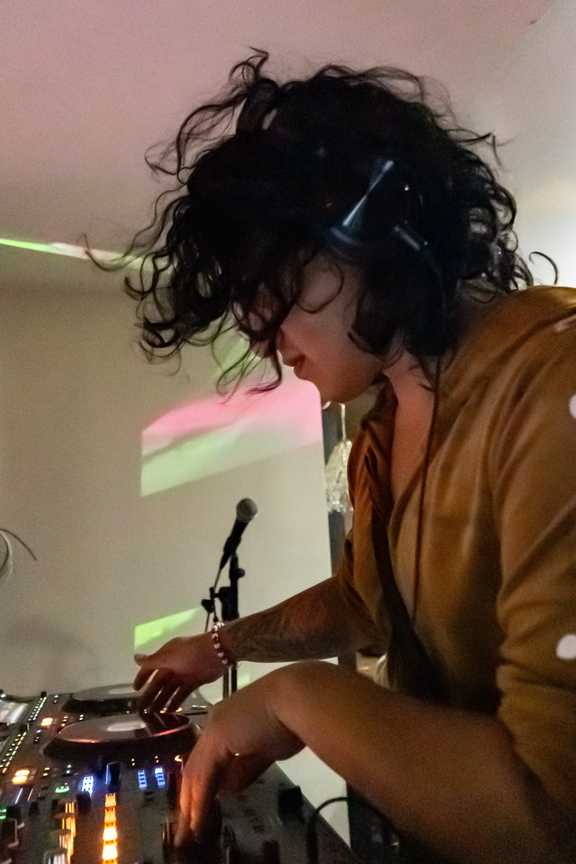 A person with curly black hair wearing headphones and a tan shirt mixes music on DJ equipment in a dimly lit room with colorful lights and a projection on the wall.