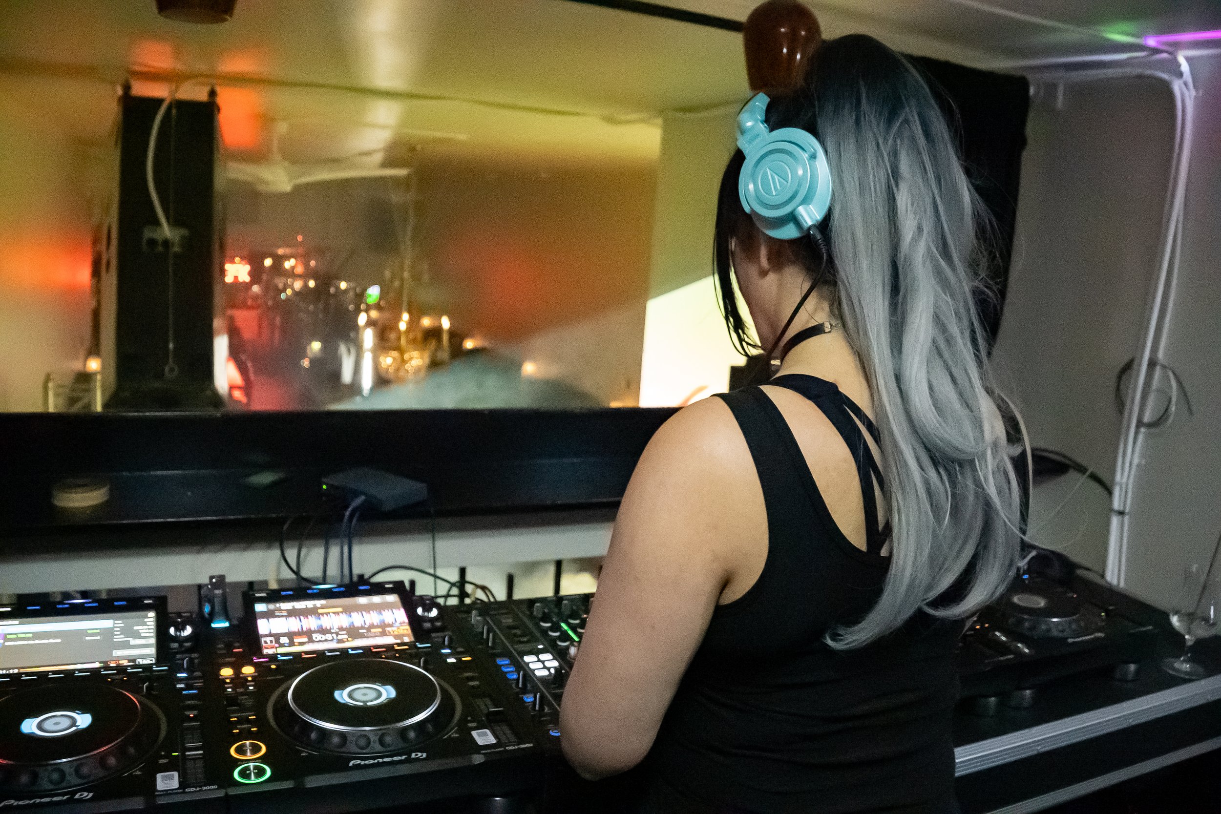 A female DJ with gray hair and black clothing wearing blue headphones, standing behind DJ equipment in a dimly lit indoor setting with colorful lights.