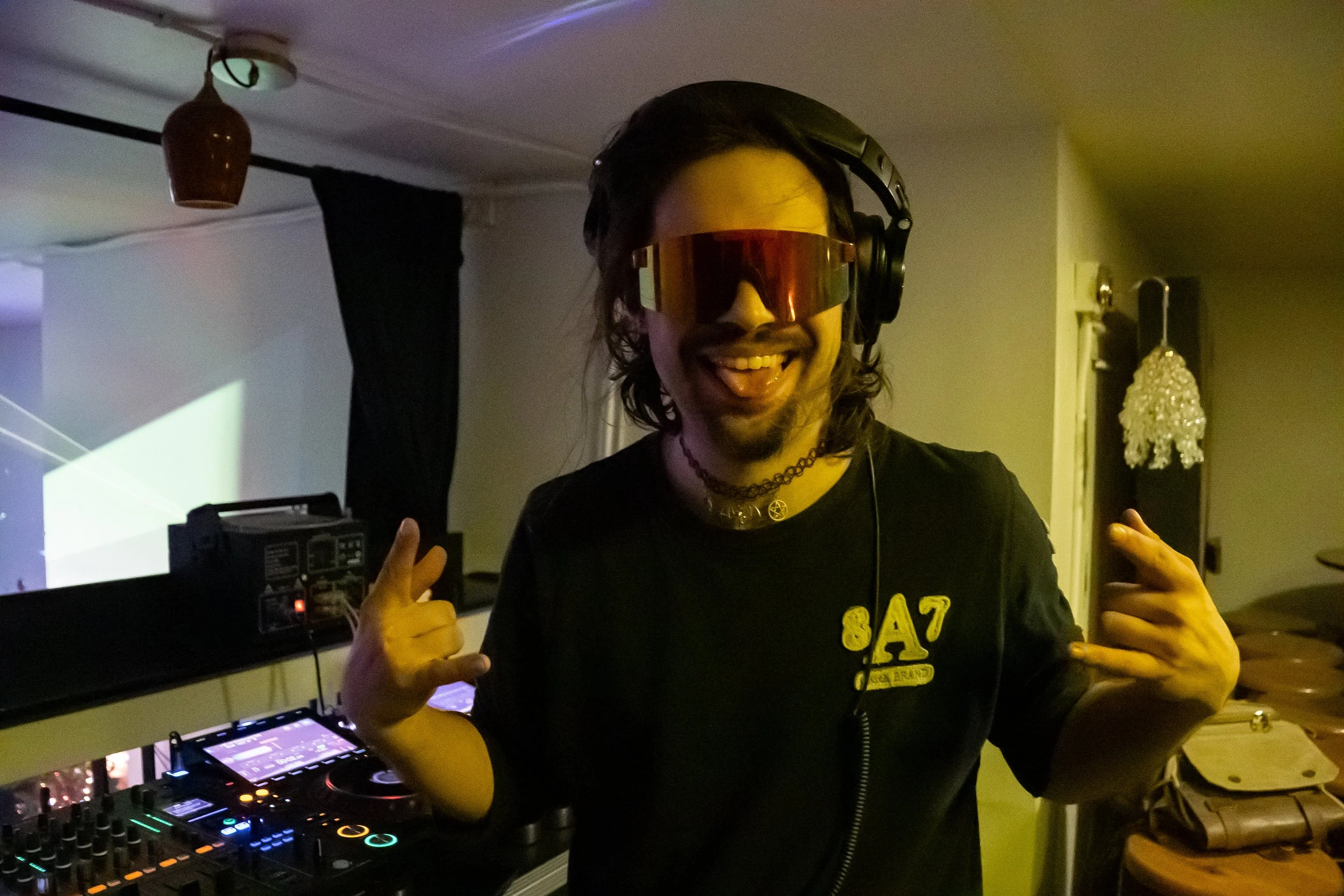 A man with long dark hair wearing sunglasses, headphones, a black T-shirt, and making rock and peace signs with his hands, standing in front of DJ equipment in a dimly lit room, smiling and sticking out his tongue.
