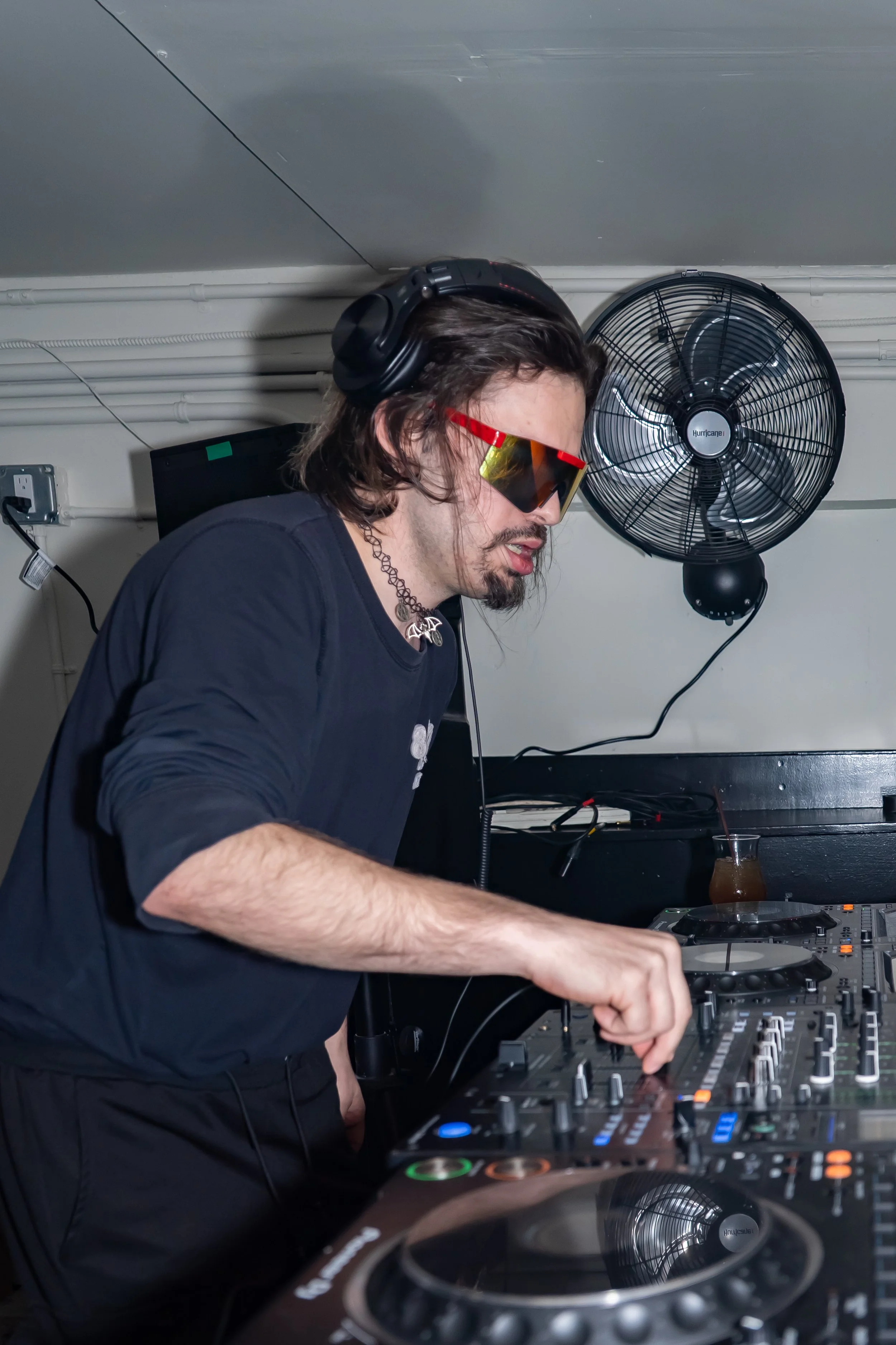 A DJ wearing headphones, sunglasses, and a black shirt is mixing music at a DJ booth in a dimly lit room with a fan mounted on the wall behind him.