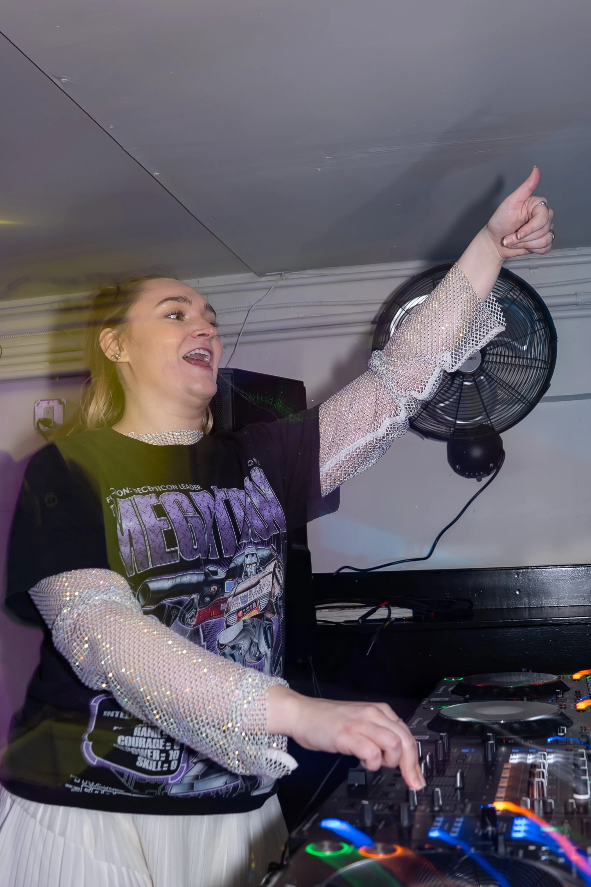 A woman DJ wearing a black "Transformers" T-shirt with silver sparkly sleeves, operating a DJ mixer with colorful lights, pointing upward with her right hand, in a room with a wall fan and electronic equipment.