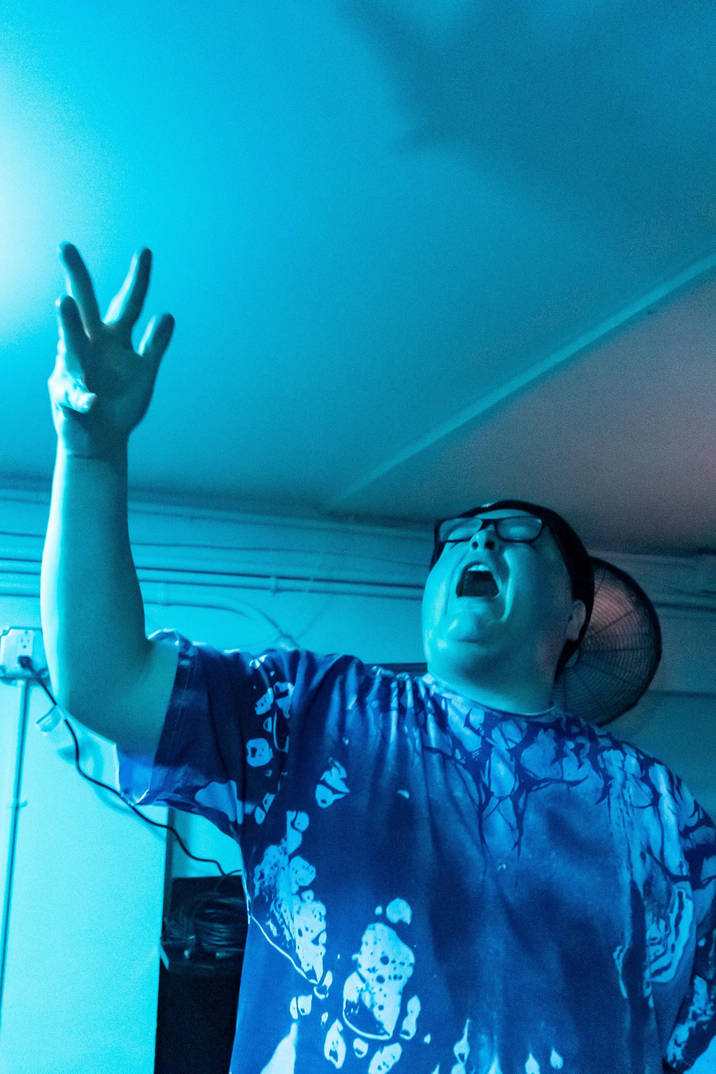 Person wearing glasses and a blue patterned shirt, with mouth open and hand raised, in a room with a fan and electrical outlets, illuminated by blue lighting.