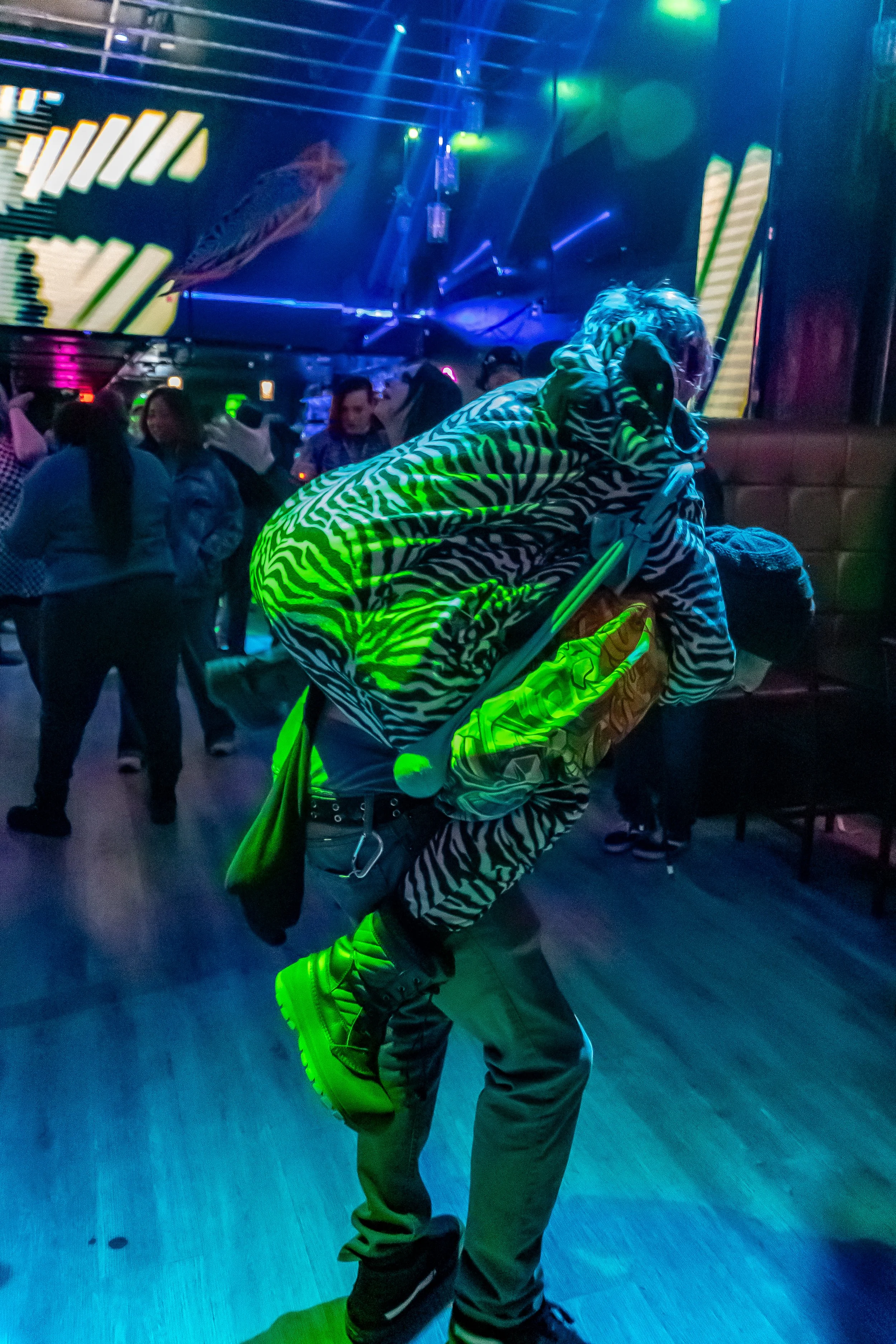 A person dressed in a zebra print hoodie and pants, with a matching mask and cap, stands in a vibrant nightclub with colorful lights, holding a smartphone.