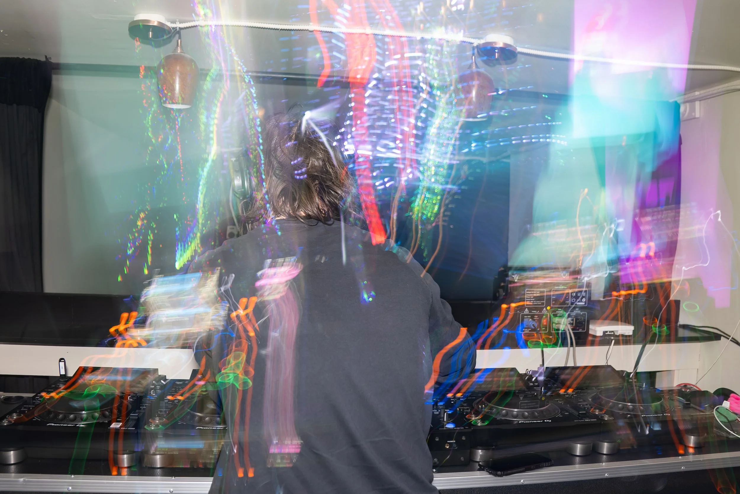 A DJ with headphones, facing away, performing at a party with colorful, dynamic light trails and blurred lights in the background.