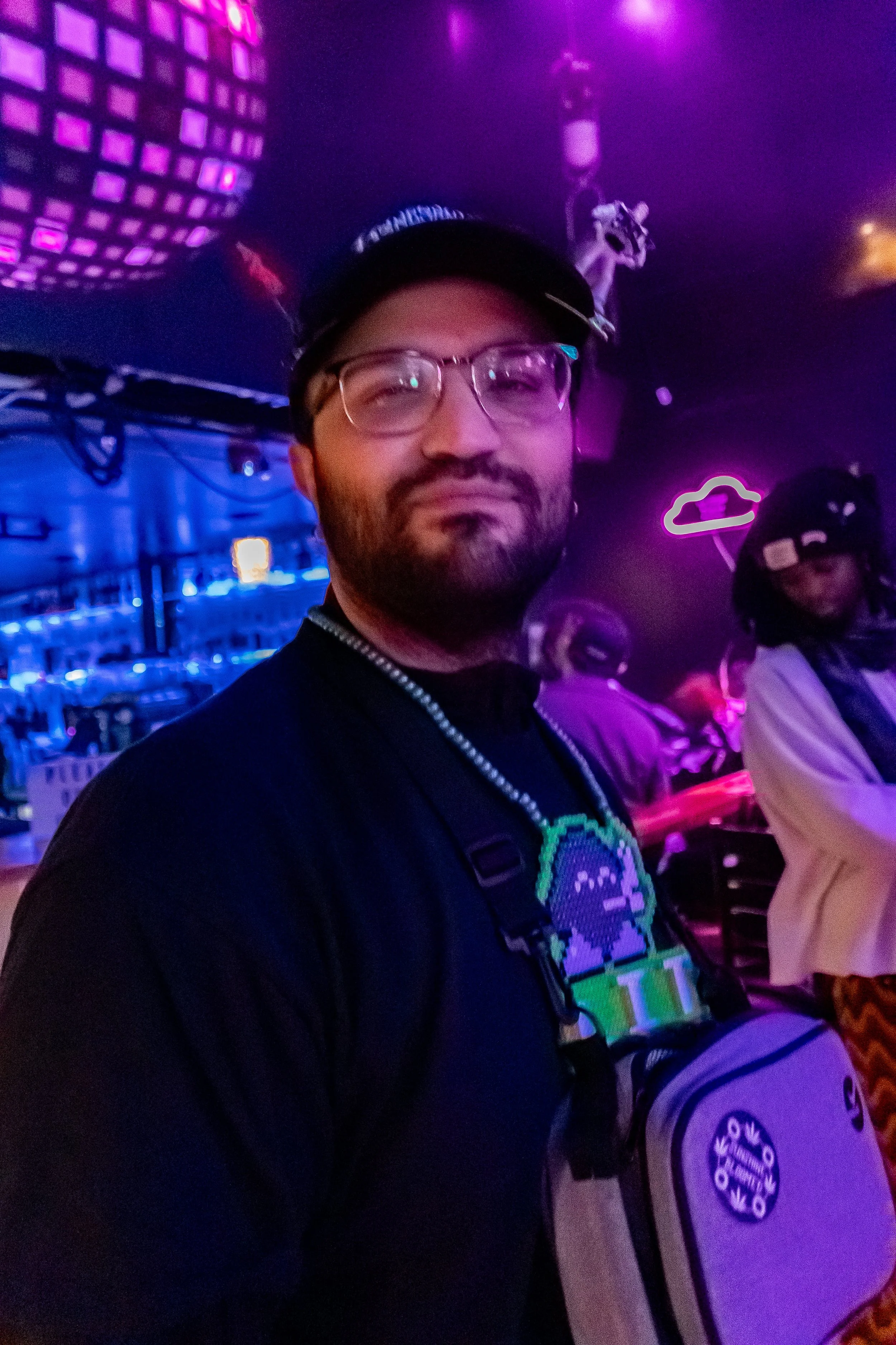 A man with glasses and a beard, wearing a black cap and a black shirt with an animated character on it, smiling in a bar with neon lights and a disco ball.