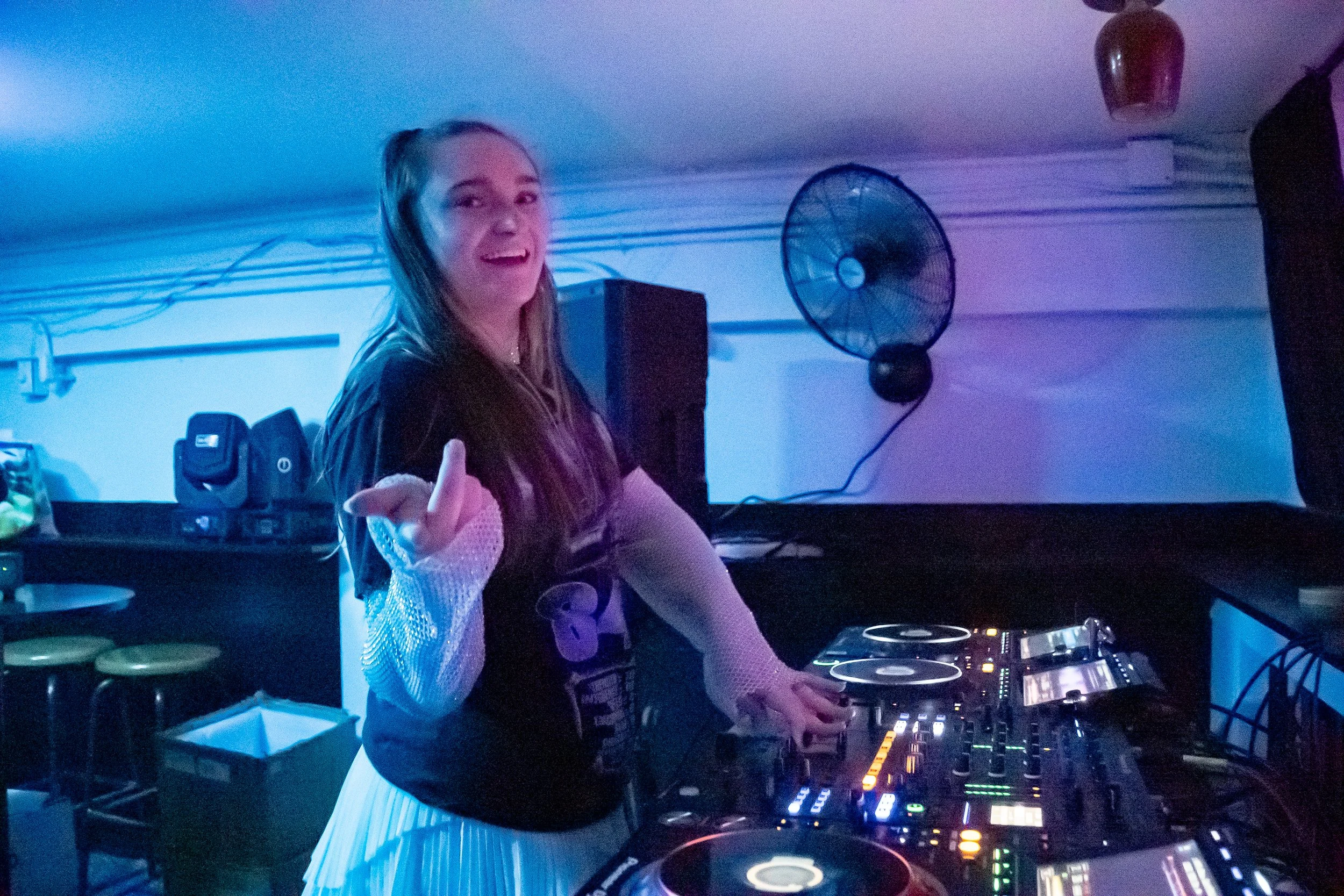 A young woman with long brown hair, wearing a black t-shirt and white mesh sleeves, stands at a DJ booth with her right hand making a gesture. She is smiling and appears to be in a dimly lit room with blue and purple lighting, with DJ equipment and a