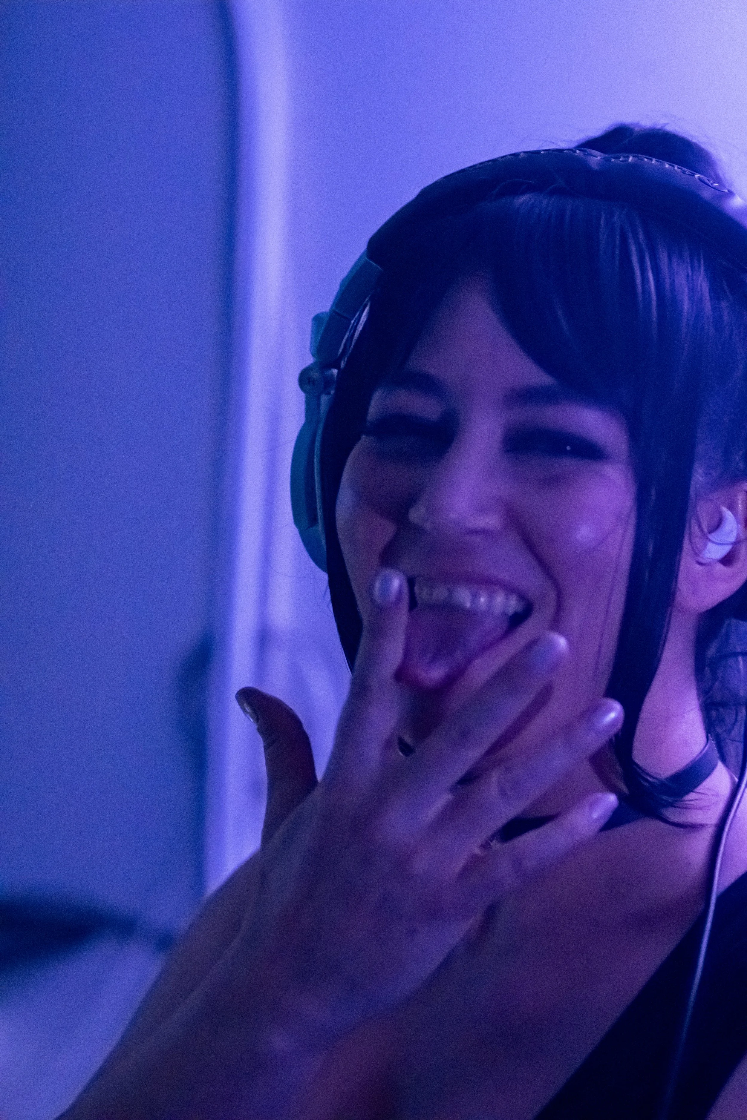 A woman with dark hair and a headset laughing with her hand near her mouth, in a blue-lit environment.