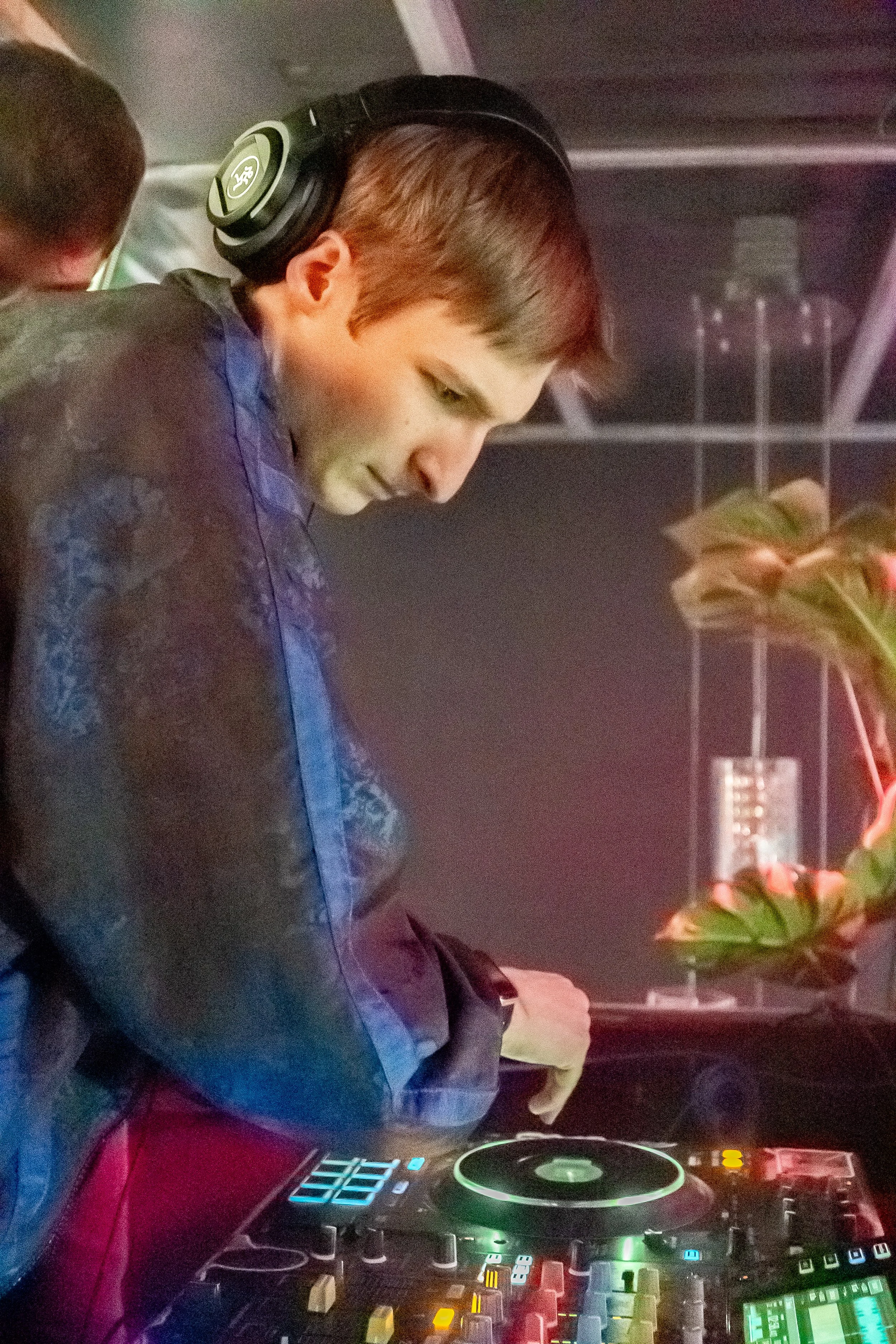 A young man with light brown hair, wearing headphones, is DJing with a mixer and turntable in front of him. He looks focused while adjusting equipment, with a plant and a black wall in the background.
