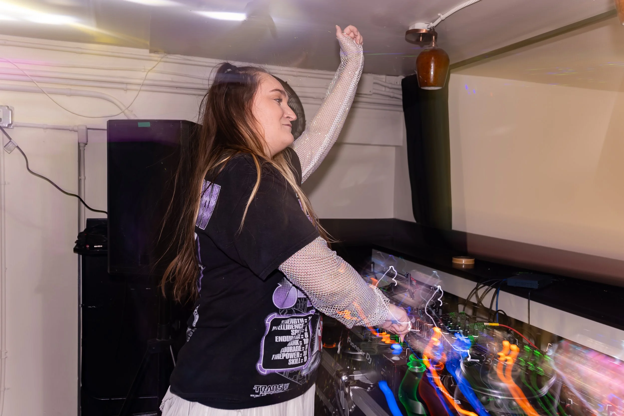 A young woman with long hair is DJing at a party, raising one arm and standing behind a DJ mixer with colorful lights.