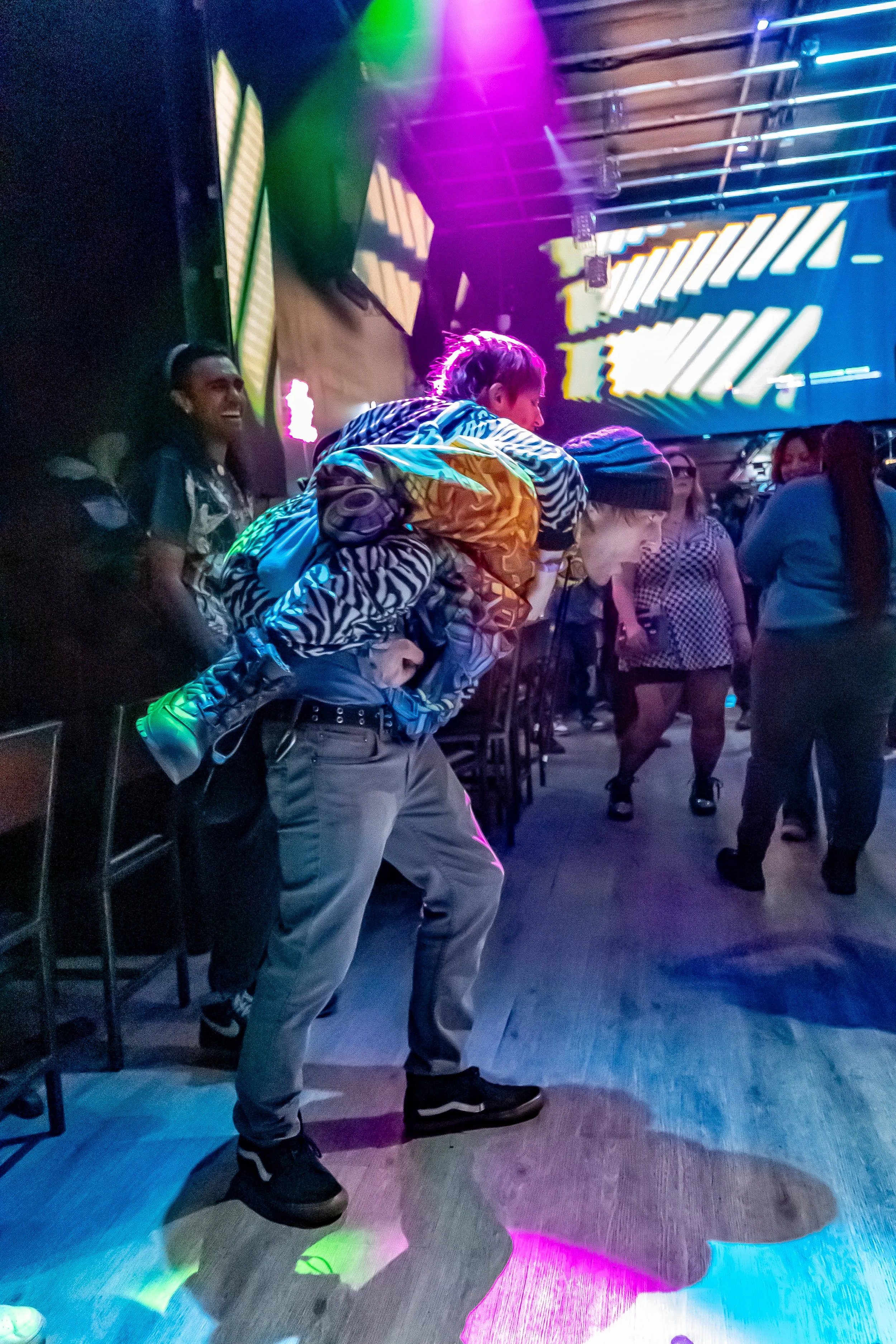 A person is giving a piggyback ride to a child at a lively nightclub or dance venue with colorful lights and a crowd of people dancing.