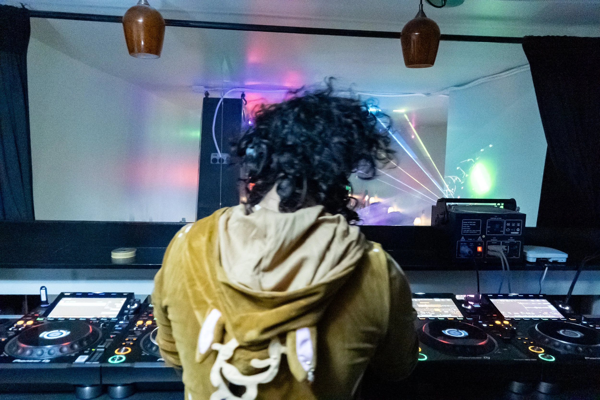 Person with curly hair wearing a yellow hoodie working as a DJ at a nightclub with colorful laser lights.