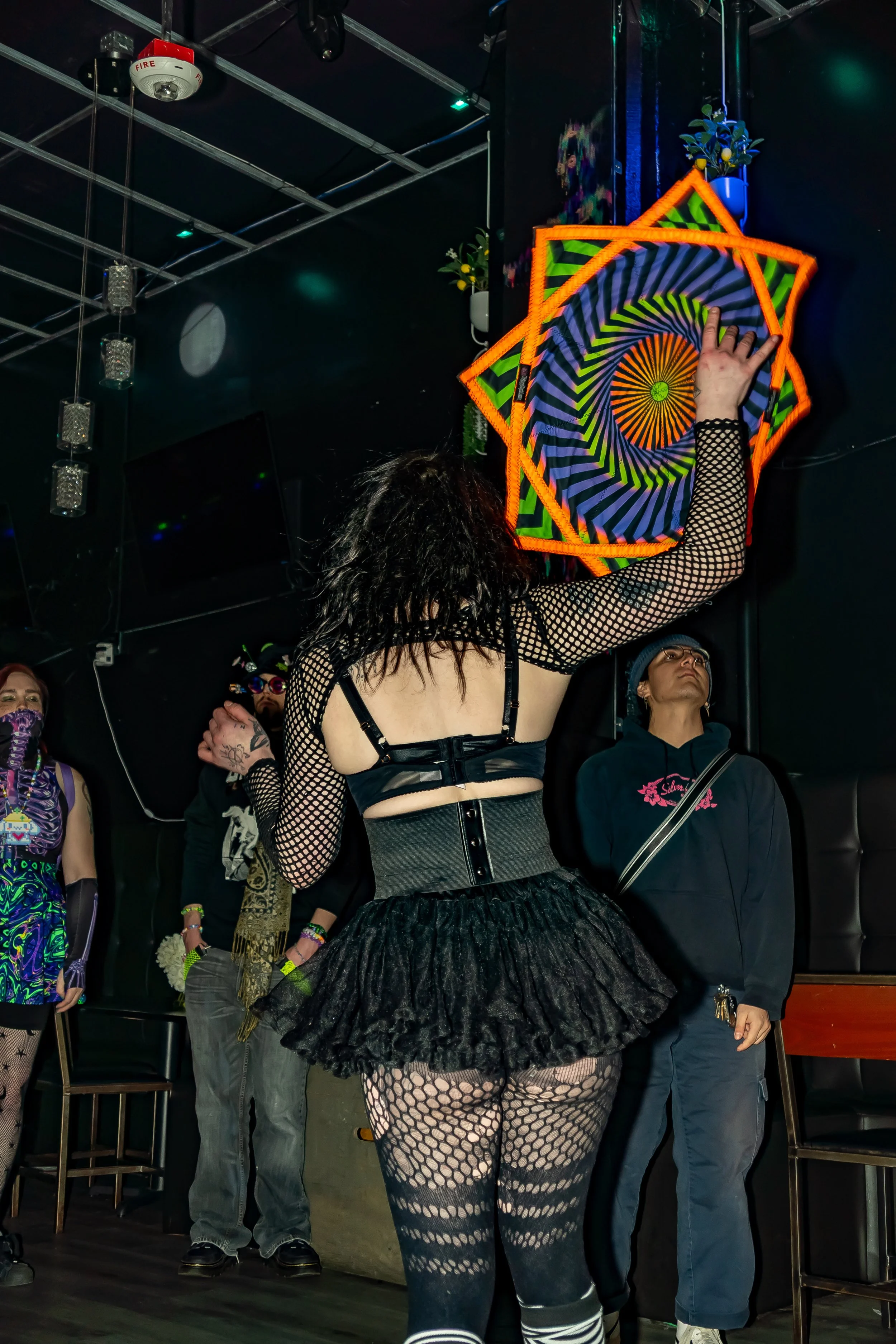 A woman with black curly hair wearing fishnet sleeves, a black corset, and a black ruffled skirt is reaching up to hang or adjust a colorful, geometric, spiral patterned decoration on a black wall. Other people are watching or waiting nearby in an in