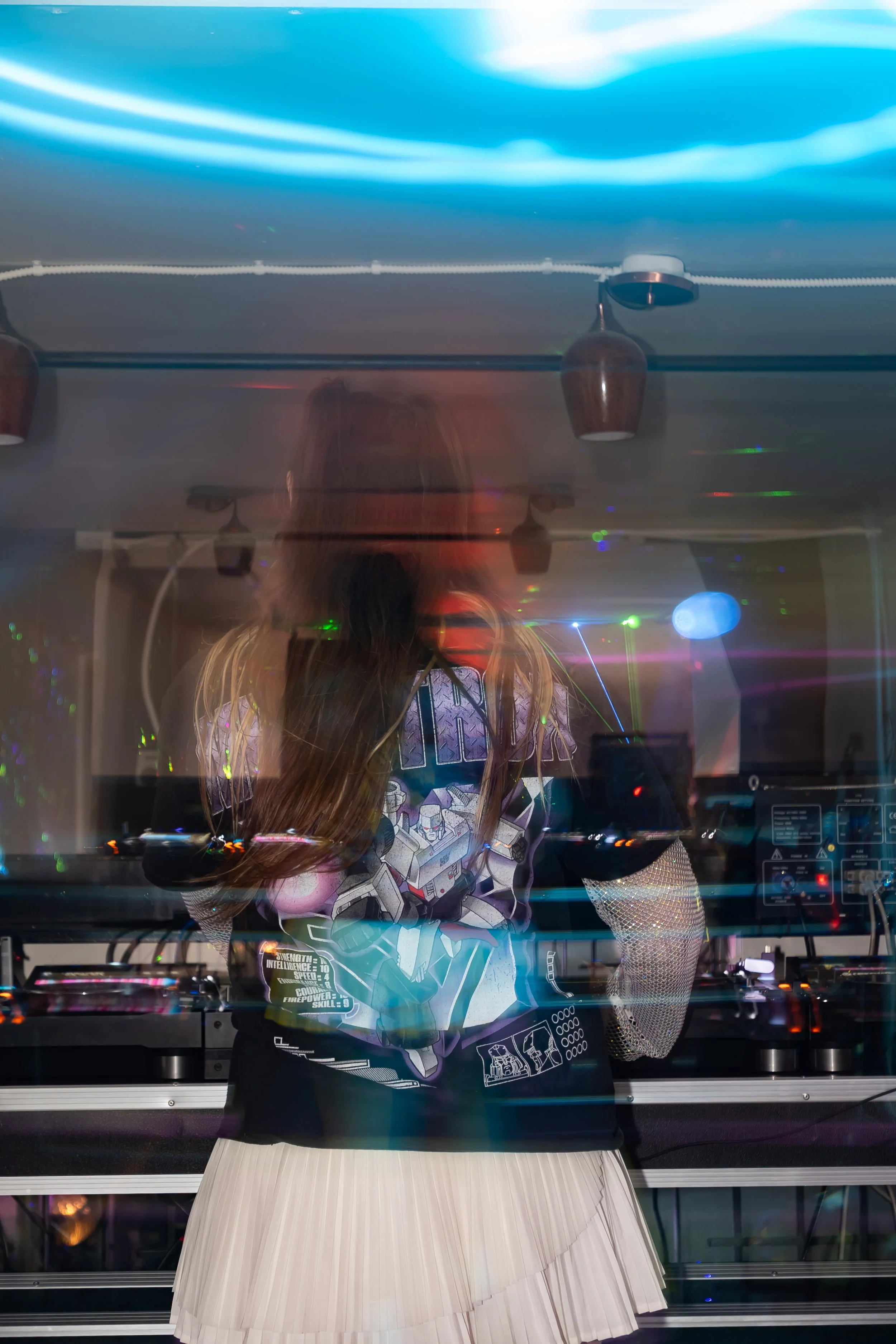 A woman with long hair wearing a graphic T-shirt and a pleated skirt, standing in front of DJ equipment with colorful lights and reflections creating a vibrant atmosphere.