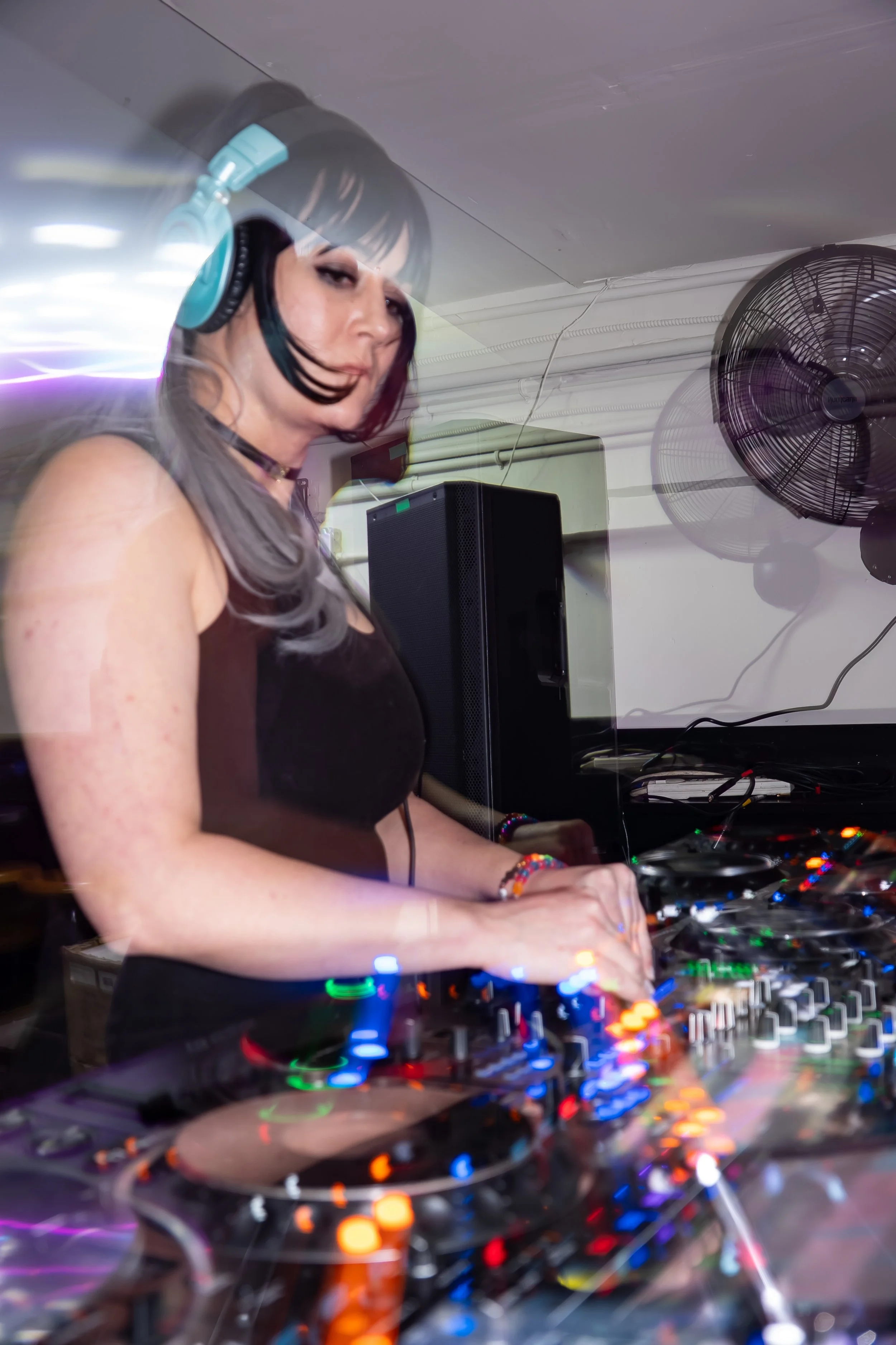 A female DJ wearing large headphones is mixing music at a club with colorful lights and sound equipment.