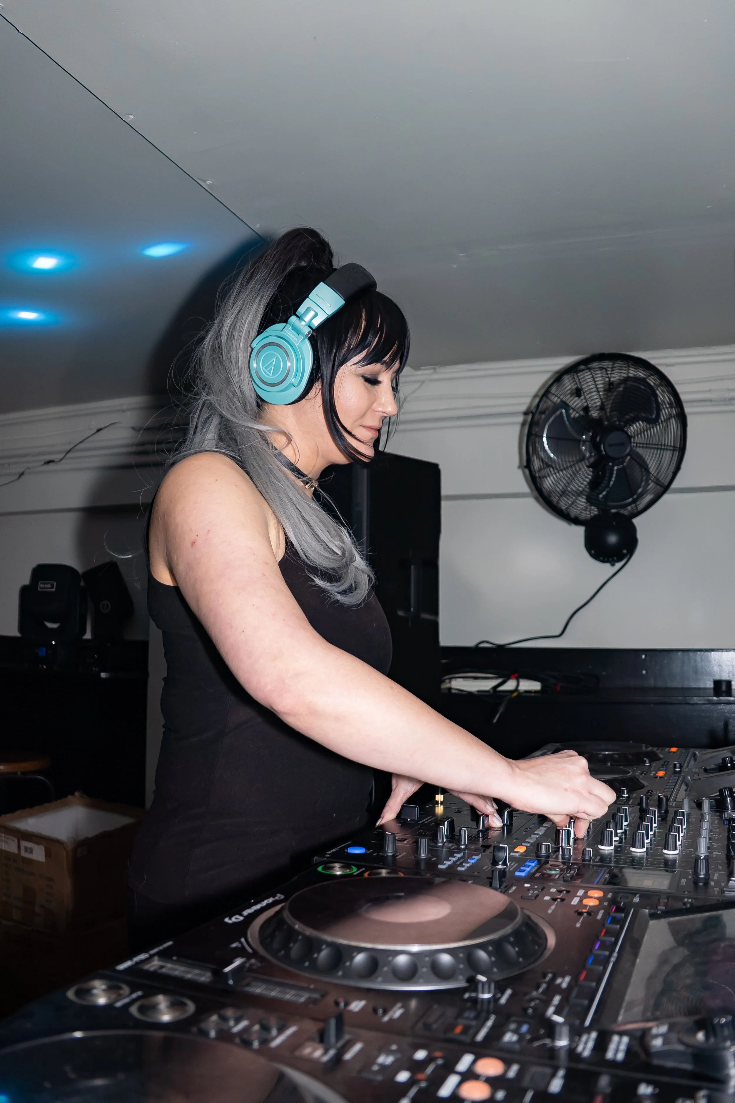A woman DJ wearing over-ear headphones is mixing music at a DJ console in an indoor setting with a wall-mounted fan behind her.