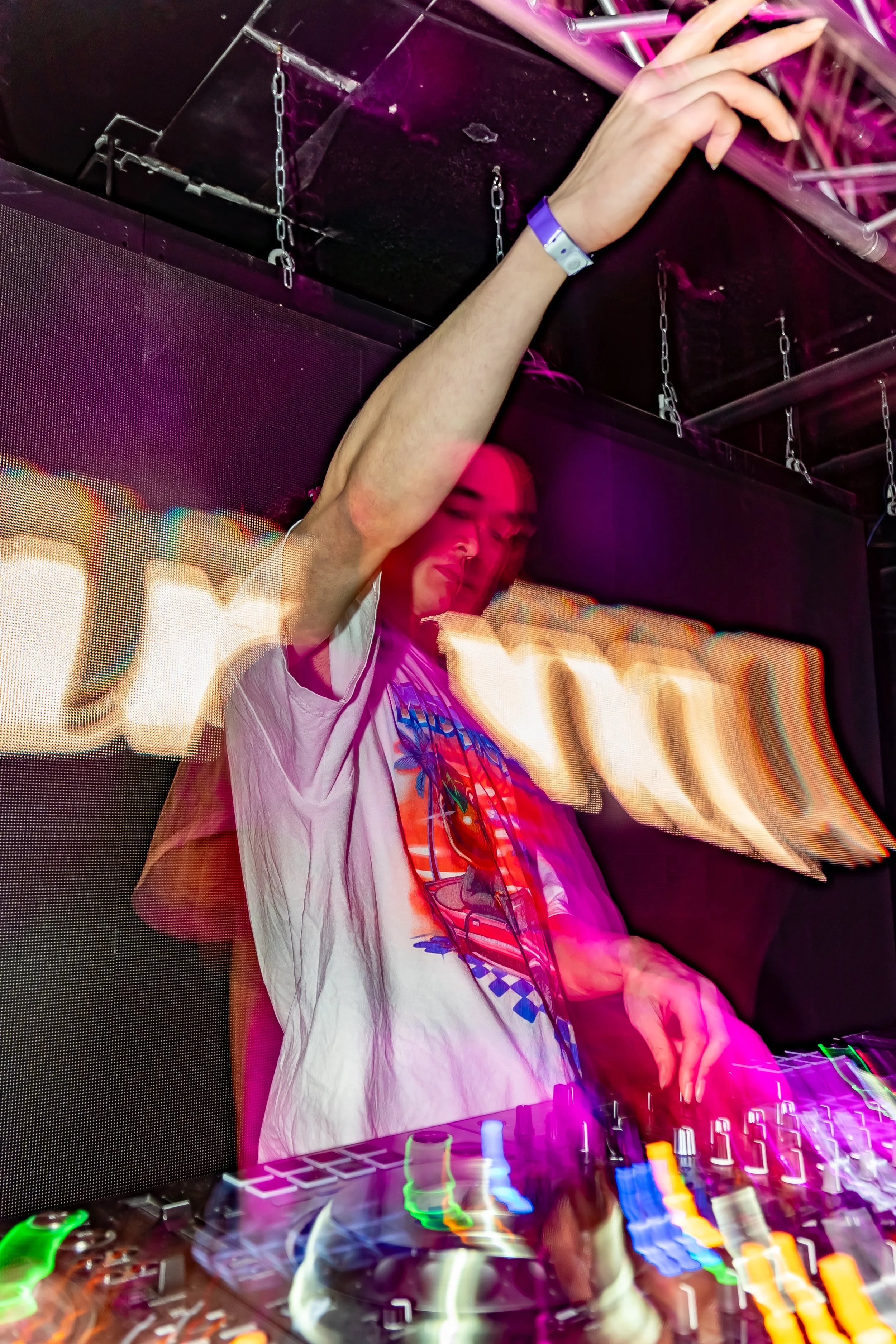 A DJ is mixing music at an indoor nightclub, with colorful lights and a digital screen in the background.
