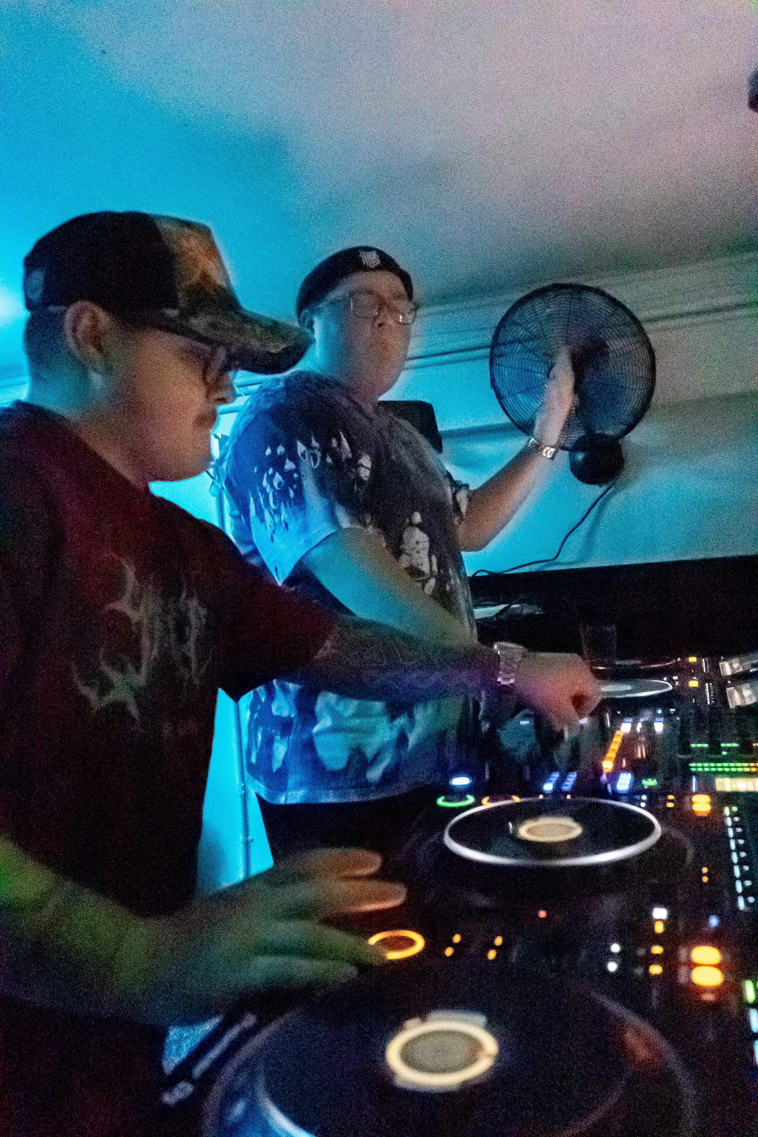 Two young men DJing at a party, with DJ equipment and a blue light illuminating the scene.