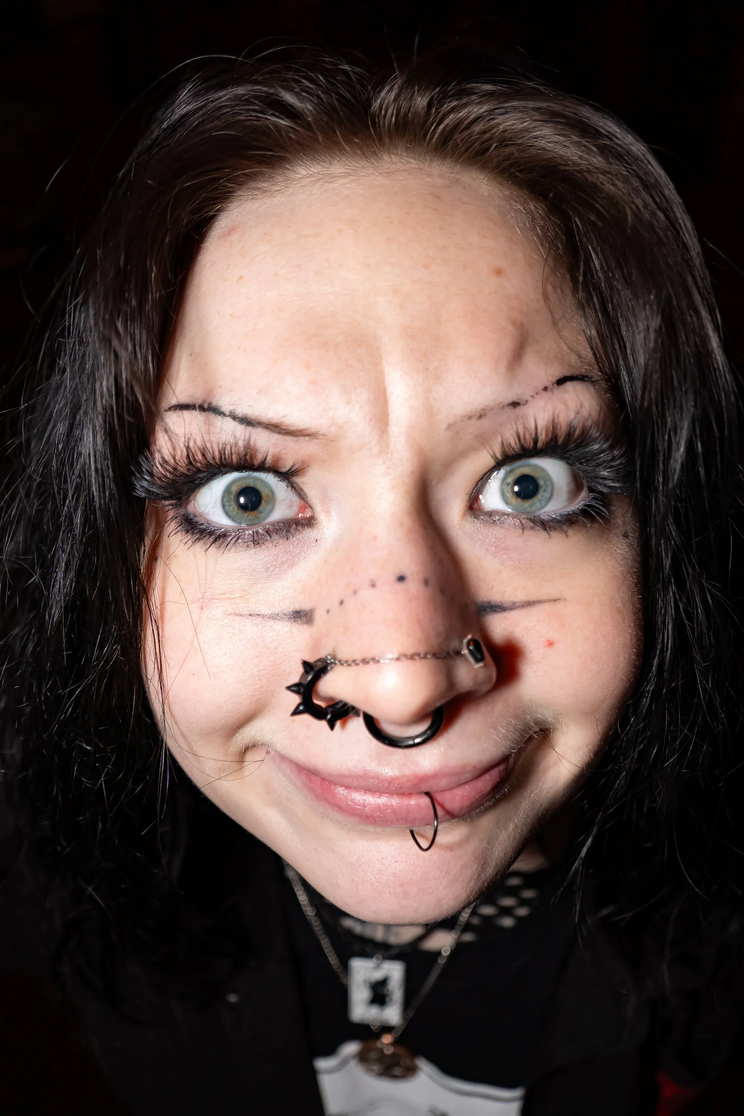 A woman with black curly hair, piercings on her nose and lips, bright green eyes, and heavy eye makeup, smiling at the camera.