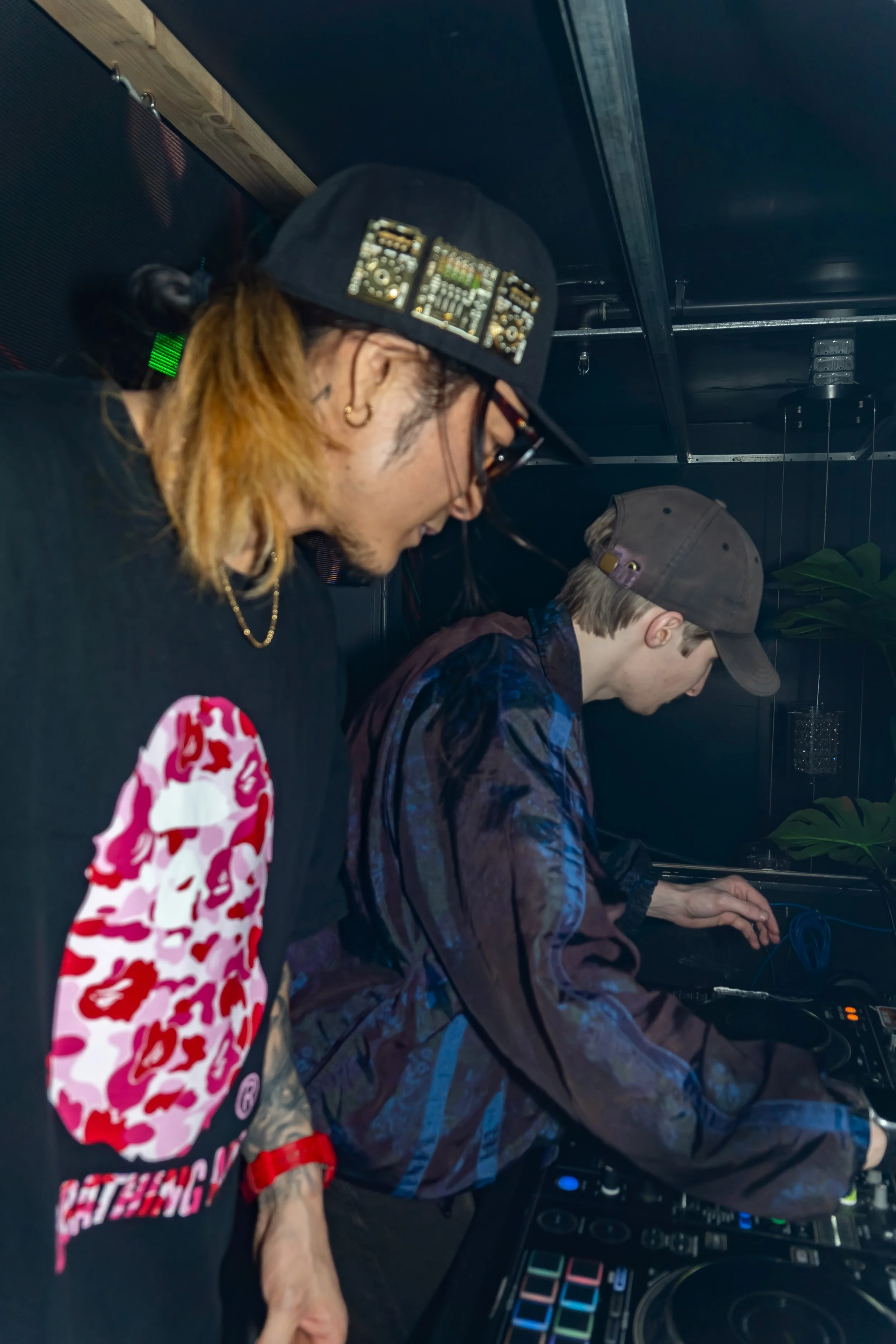 Two men, possibly DJs, are working at a DJ booth with equipment, one wearing a black hat with Technics turntables on it, and the other wearing a baseball cap, in a dark room with some plants visible.