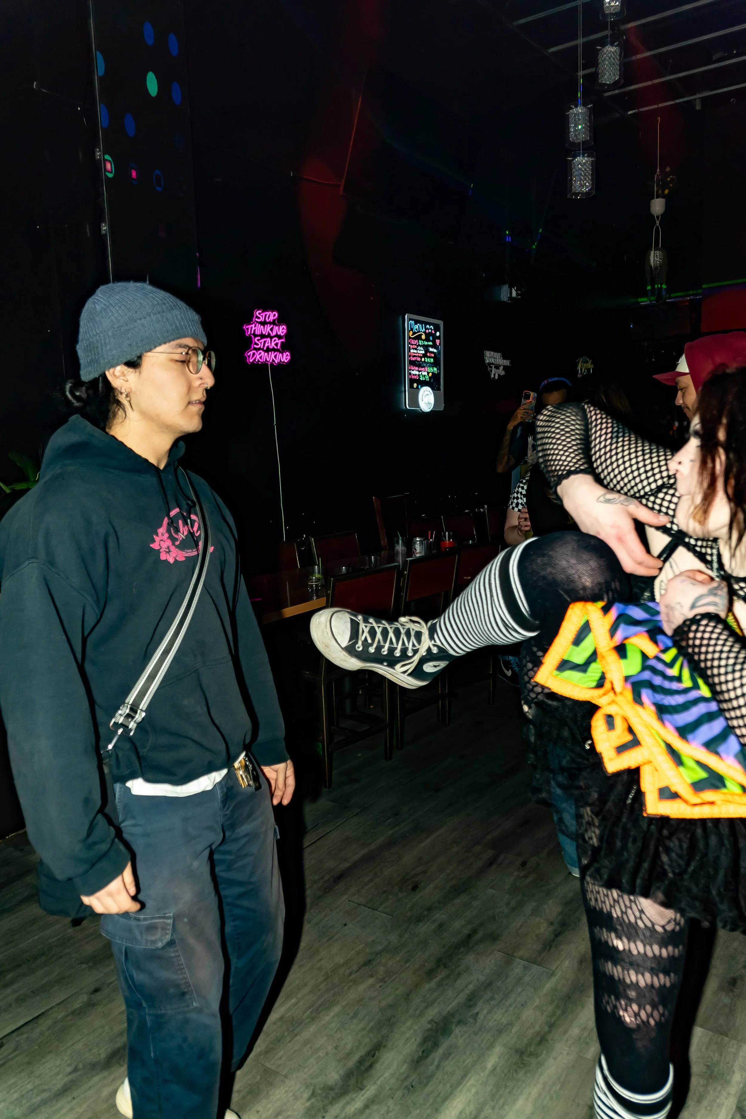 Group of people in a dimly lit room with neon lights, one person sitting on another's lap, others standing nearby, and neon signs on the wall, including one that reads 'Stop Thinking Start Drinking'.