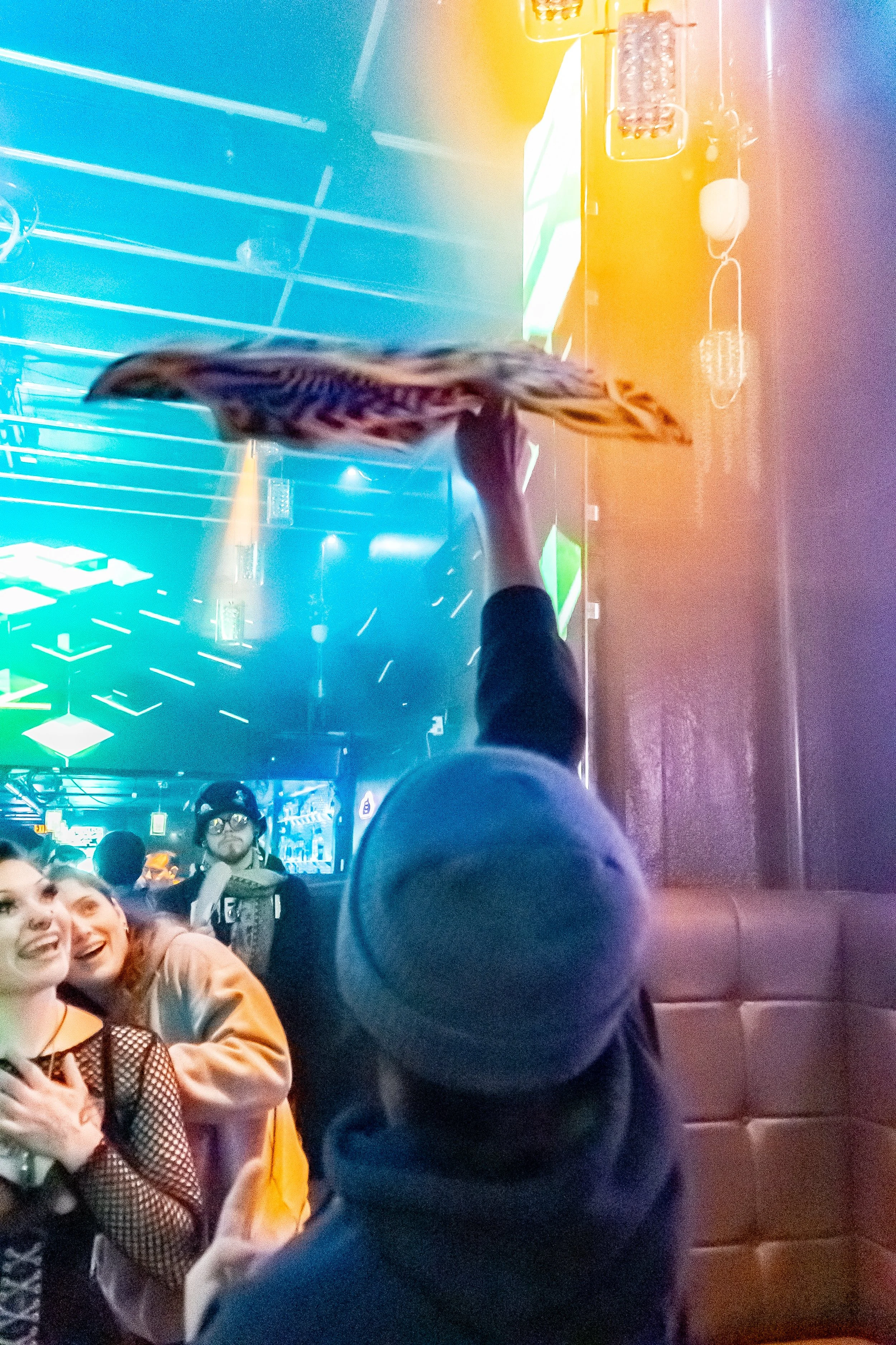People at a nightclub, one person balancing a shoe on their finger, others smiling and enjoying the lively atmosphere with colorful neon lights.