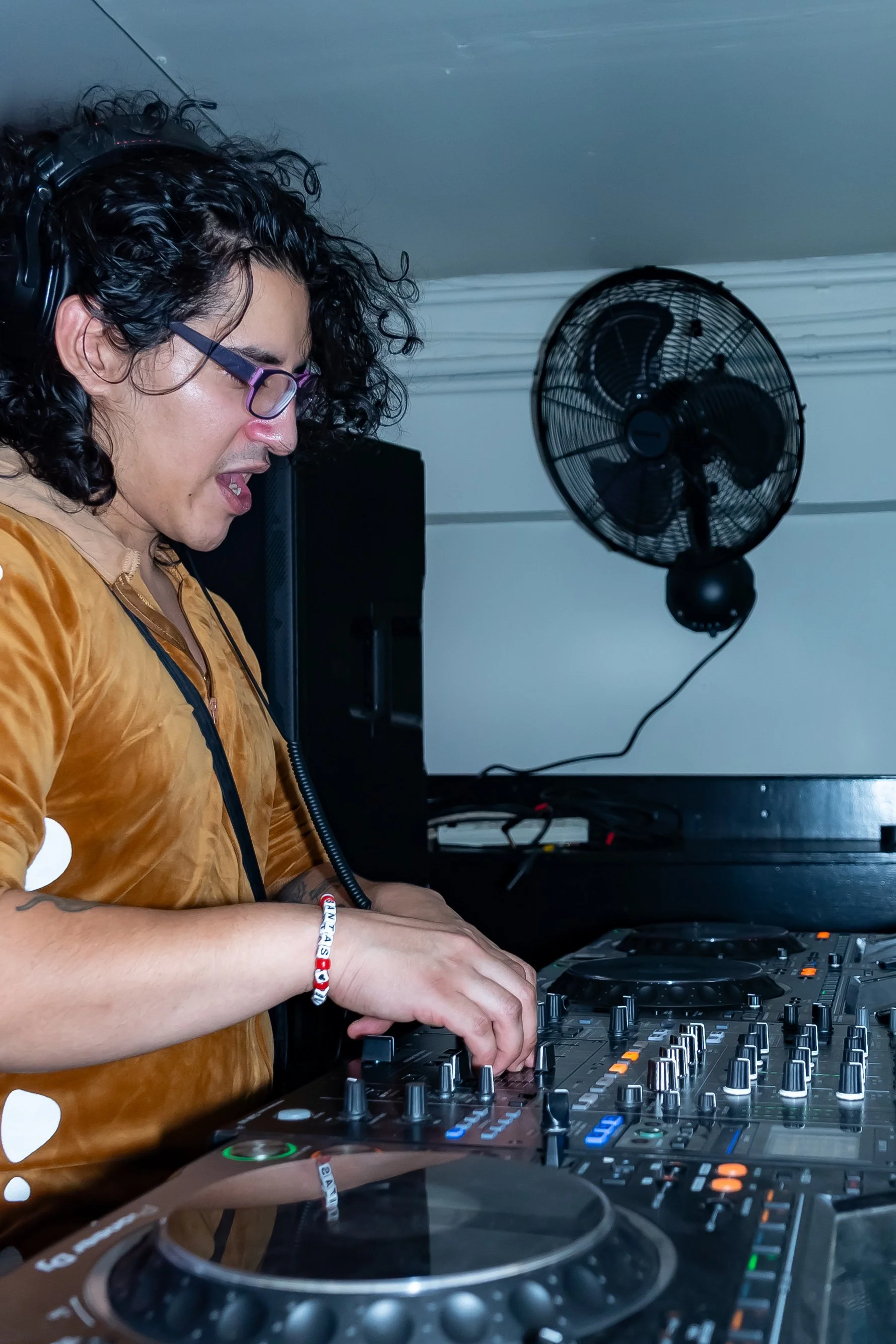 DJ with curly dark hair, glasses, and a tan hoodie, wearing headphones, operating a DJ controller in a room with a wall-mounted fan in the background.