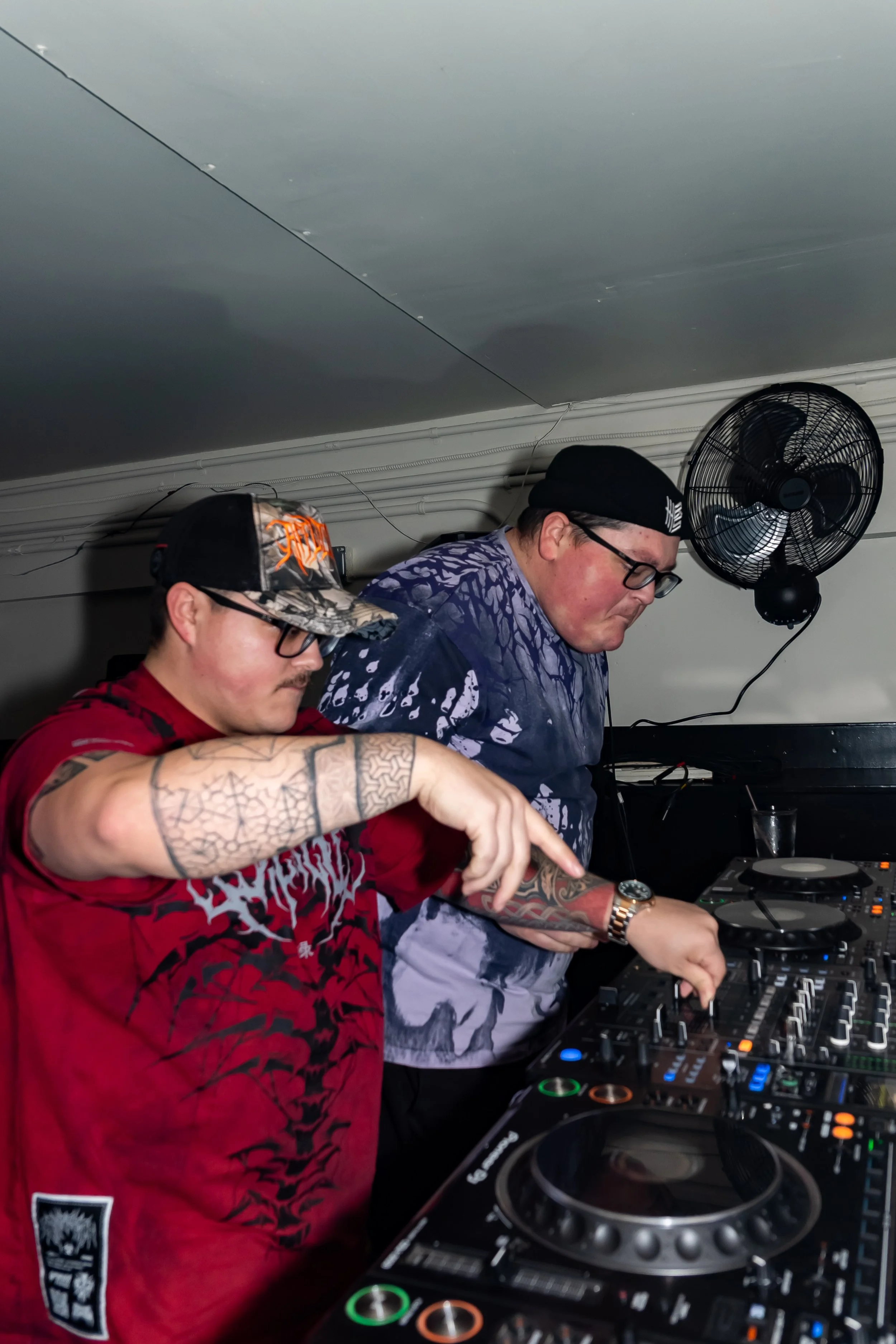 Two men DJing at a club, using professional DJ equipment, one is wearing a red shirt and a camo cap, the other is wearing a tie-dye shirt and a black beanie installed on the ceiling, with a wall fan in the background.